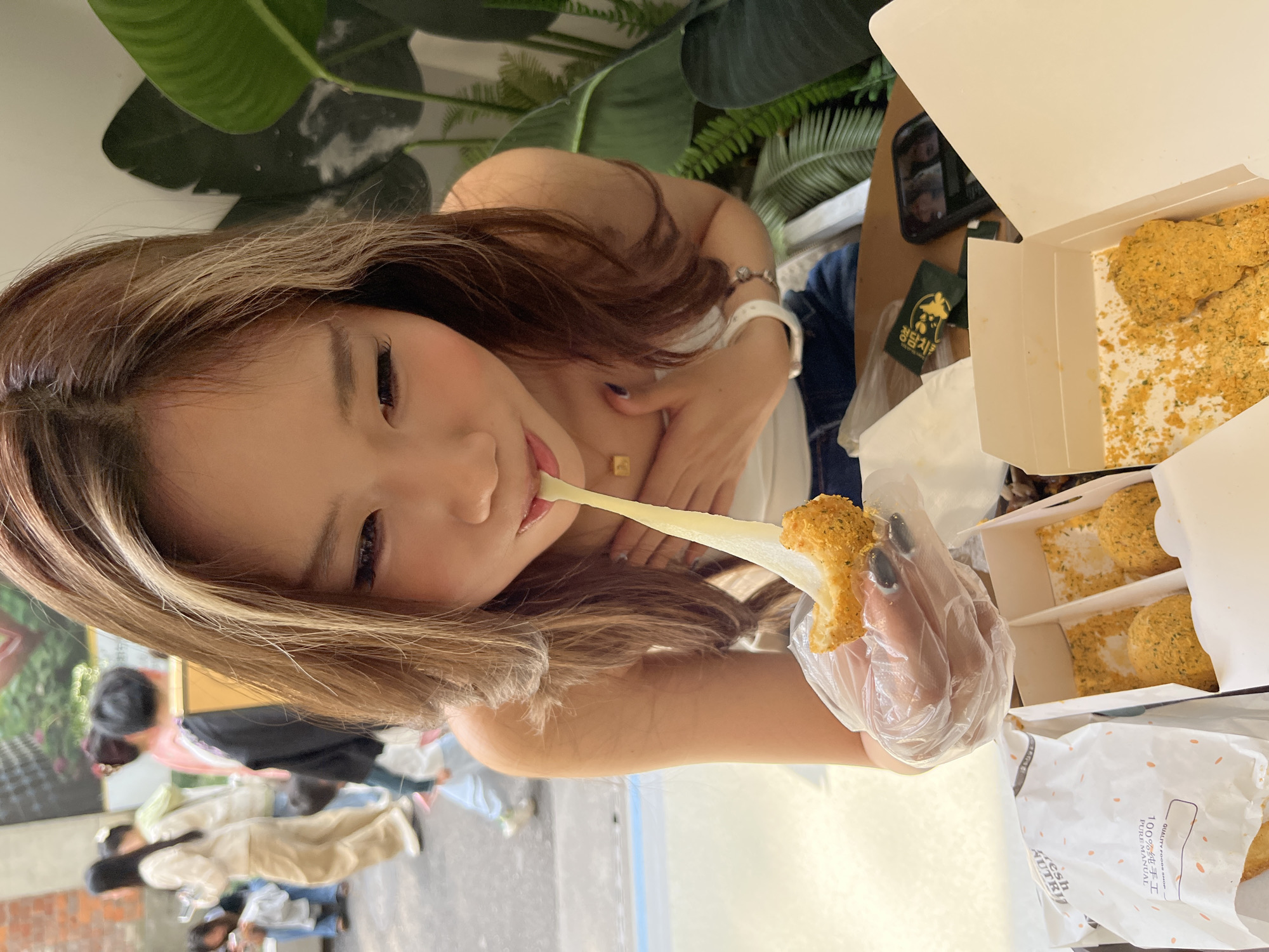 An eye-level, medium indoor shot shows a woman in Chenghua District, China, eating cheese-coated fried snacks. The woman is turned with her face left of center. She has brown hair with lighter highlights, fair skin, and is pulling a strand of melted cheese out of her mouth with one hand, while the other hand, which is in a plastic disposable glove, holds a cheese-covered fried snack. She is wearing a white top, has a bracelet, and a gold square necklace. In front of her are two white takeout boxes containing cheese-covered fried snacks, as well as a white bag with the snacks as well. A green plant and a dark item are partially visible at the top of the frame. In the background, out of focus, are people walking outside. The ground appears to be concrete.