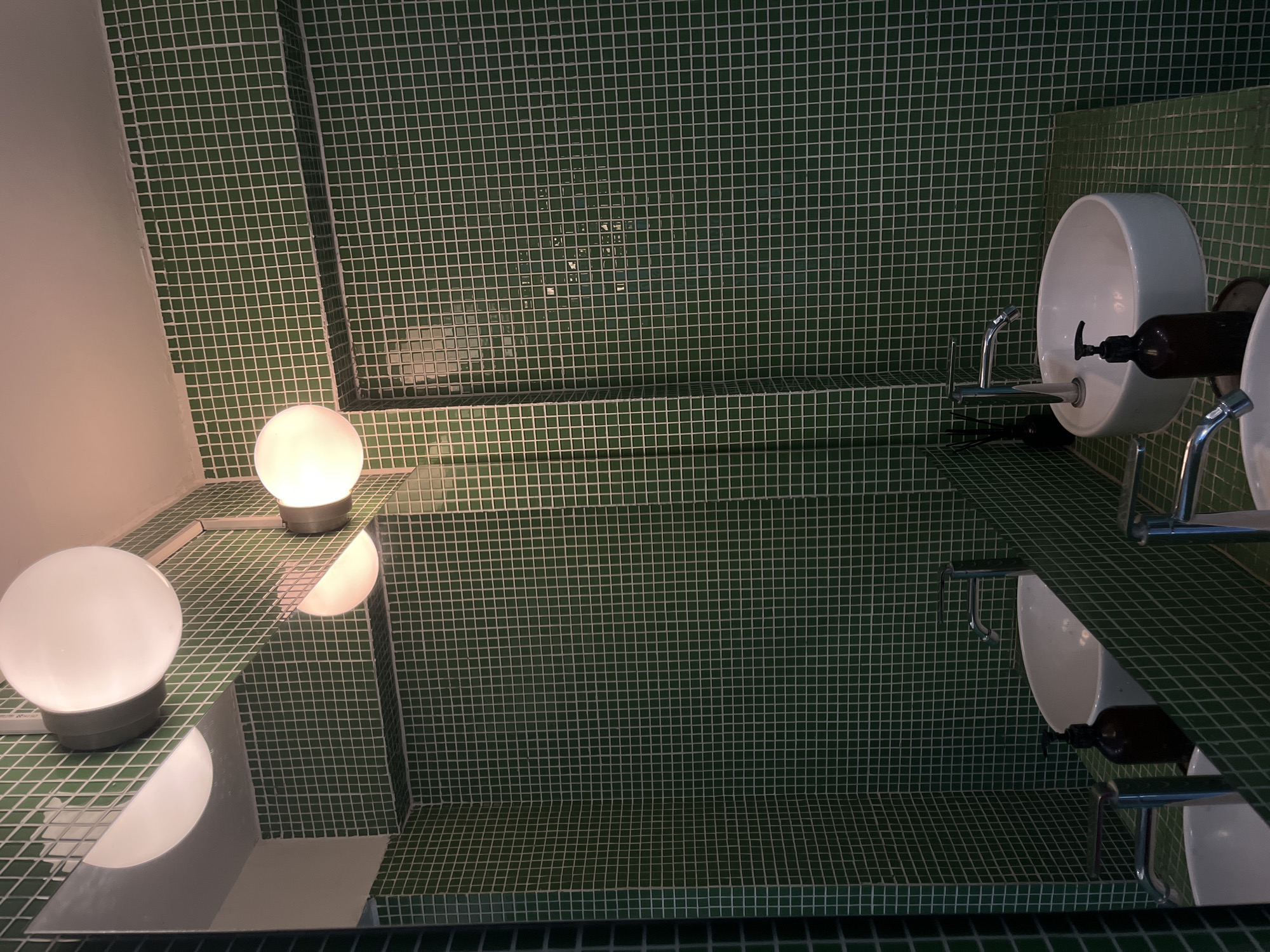 The image depicts a bathroom interior in North Kuta, Indonesia. The dominant feature is the green, square-tiled walls covering the room. The shot is taken looking into a mirror, thus doubling the visual information. Two spherical light fixtures are visible on the left, one on the counter and the other in the reflection. A white, bowl-shaped sink with chrome fixtures sits to the right, accompanied by a dark-colored soap dispenser. The sink and its fittings are also mirrored.