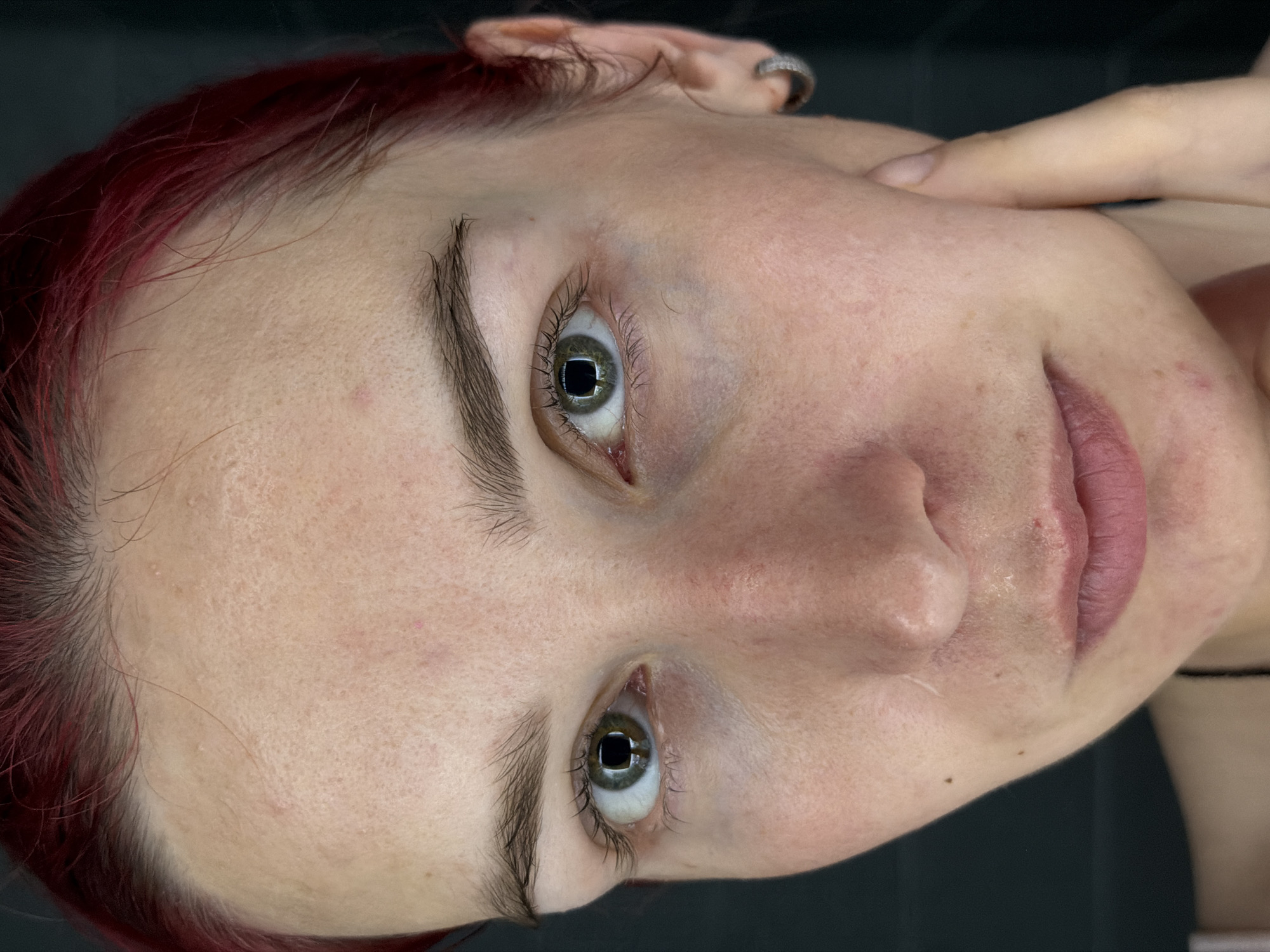 A close-up, frontal portrait of a young woman with red hair, her face filling the frame. Her eyes are wide open and looking directly at the camera, with green irises and dark pupils that reflect the light source. Her skin appears to have some texture, with visible pores and a few small blemishes. There are faint lines around her eyes and mouth, and some redness on her cheeks. Her lips are closed and painted a muted pink. Her eyebrows are dark and well-defined. She is wearing a small, silver hoop earring in her visible right ear. Her right hand is raised, with her index finger and thumb gently touching her cheekbone. The background is dark and out of focus, providing no environmental context. The image is sharp and well-lit, highlighting the details of her facial features. The location of Chaikovsky, Russia is not discernible from the image.