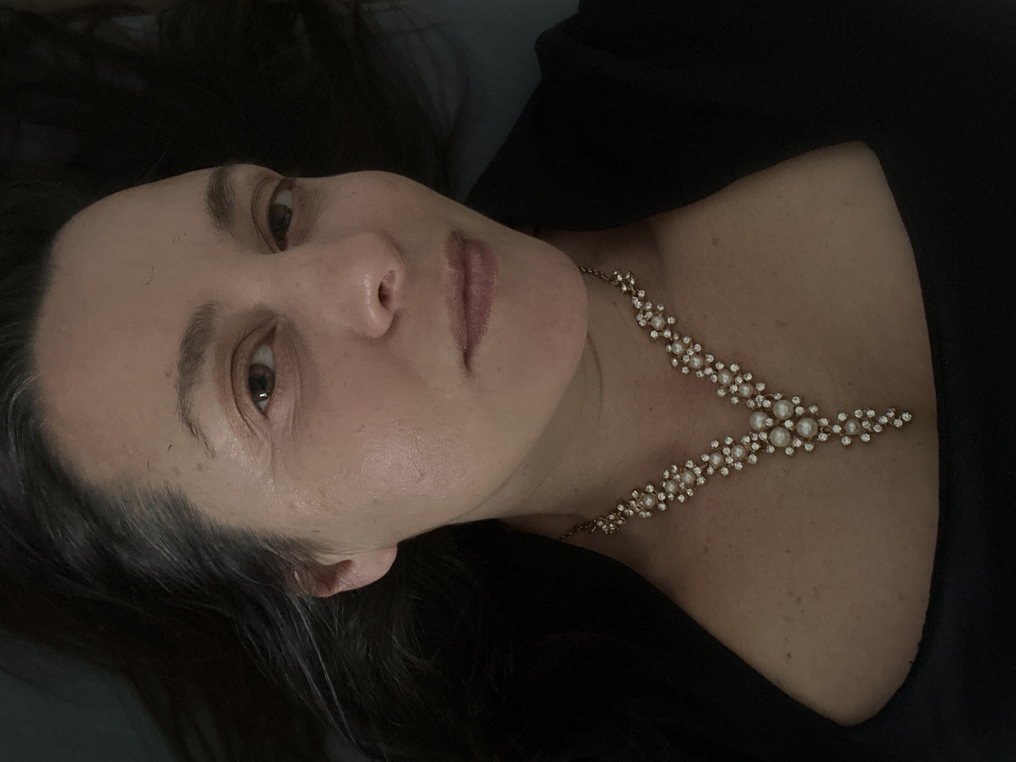 A person, likely female, is depicted in a close-up portrait, reclining with her head slightly tilted. Her dark, long hair is spread around her head, contrasting with her fair to medium complexion. Her gaze is directed towards the viewer, with a neutral to slightly contemplative expression. Her eyebrows are dark, and her lips are softly colored and slightly parted.
She is adorned with an elegant gold-toned necklace featuring an intricate design of small white pearls and sparkling clear stones, arranged in a floral or snowflake-like pattern. The necklace forms a 'V' shape, resting gracefully on her décolletage. She is wearing a simple black garment, possibly a top or dress, visible on her shoulders and upper chest, providing a dark backdrop that highlights the jewelry.
The lighting is soft and appears to be artificial, illuminating her face and the necklace while the background remains dark and indistinct, suggesting an indoor setting. The image focuses on the individual and her prominent accessory, captured in a moment of repose. No specific activity beyond posing for the photograph is evident. There is no visible text. The location context for this image is Riga, Latvia.
