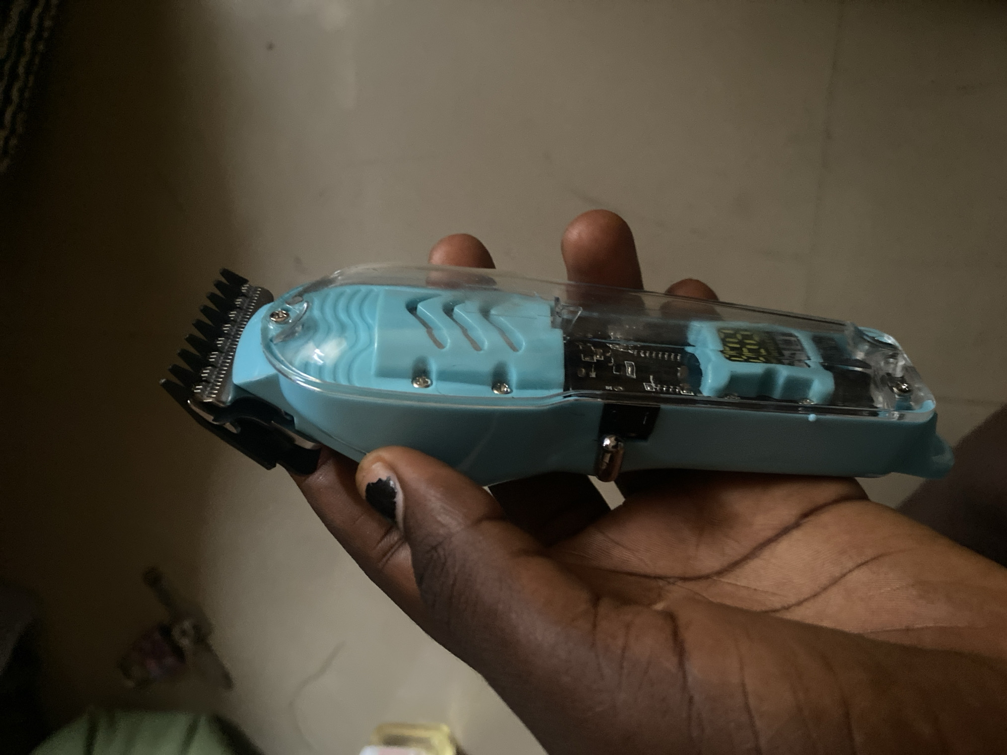 The image shows a close-up of a hand holding a light blue and clear cordless hair clipper. The clipper has a translucent top, revealing internal electronic components and a small digital display. The cutting head of the clipper, made of metal teeth, is visible on the left side. The hand holding the clipper has dark skin and has a black nail polish on one finger. The background is out of focus and appears to be an indoor setting with a textured wall and floor. The lighting suggests it could be daytime, but it's difficult to determine the exact time. There is no specific activity or event taking place beyond the presentation of the hair clipper. No discernible text is visible on the clipper or in the background. The overall impression is a simple product shot.
