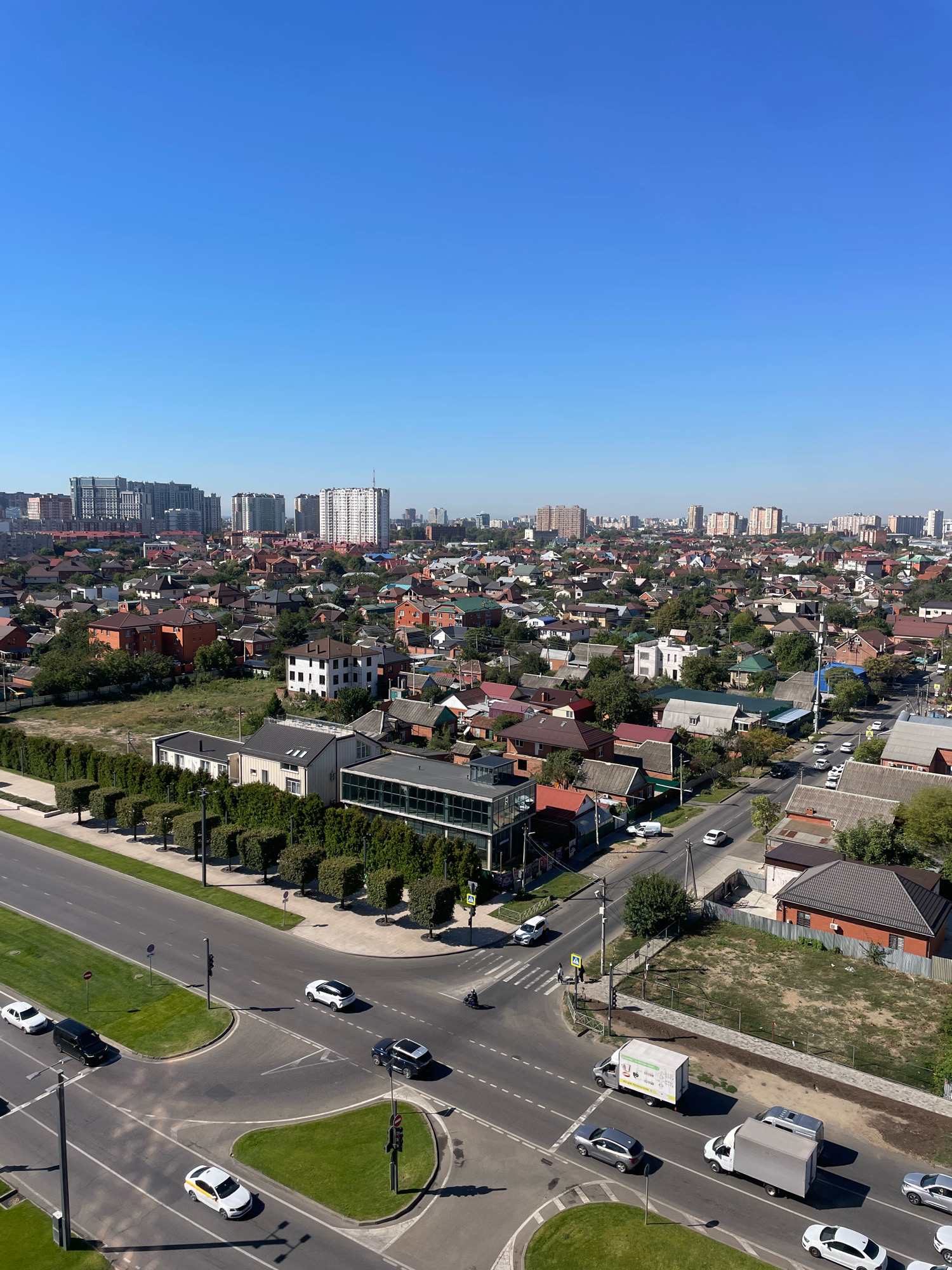 This is an aerial, high-angle, full shot of a city intersection in Krasnodar, Russia. The sky is clear and blue. The scene depicts a mix of residential buildings with red-tiled roofs and modern, multi-story buildings in the distance. A paved road network is visible, with several cars and trucks driving on it, including a white truck with graphics and a large white box truck. There are also traffic lights, streetlights, and green grassy areas. The residential buildings are primarily single-family homes. The modern buildings in the background suggest urban development.