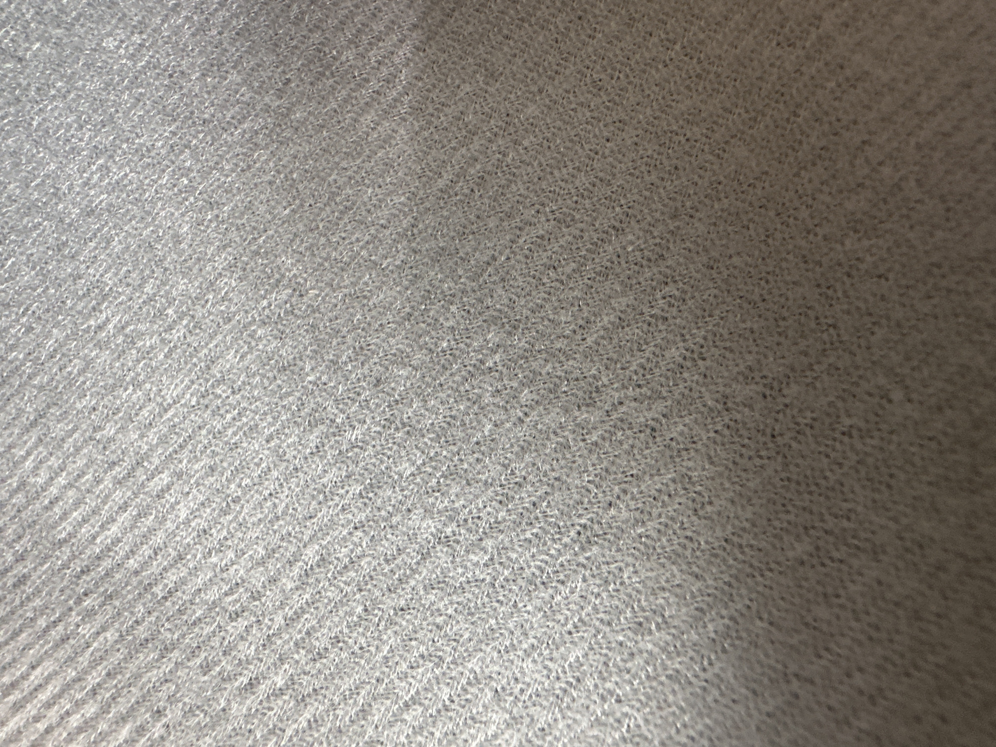 This is a close-up, abstract photograph of a textured, gray fabric. The fabric appears to be woven, with a fine, fuzzy texture. In the lower left quadrant, there are distinct diagonal lines suggesting a ribbed pattern. The lighting creates a gradient across the surface, with the left side being brighter and more detailed, while the right side is darker and slightly out of focus, indicating a light source from the upper left. There are no people, objects, or recognizable scenes visible. The image focuses solely on the tactile qualities and visual pattern of the material. No text is visible.