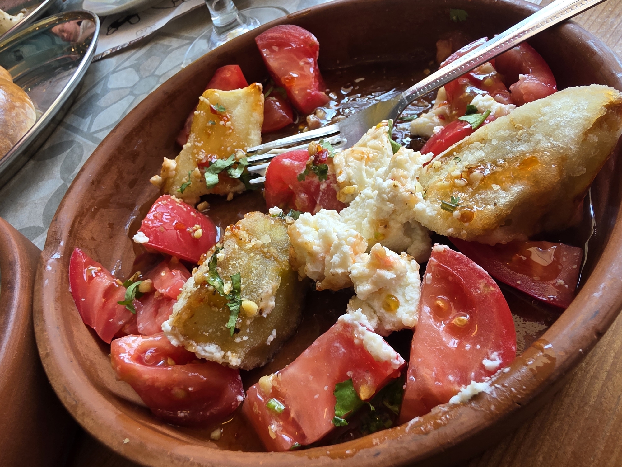 A close-up, overhead shot shows a rustic, earthenware bowl filled with a colorful salad. The salad features sliced red tomatoes, creamy white cheese, and pieces of what appear to be fried eggplant or zucchini, all drizzled with a glistening sauce and sprinkled with chopped green herbs. A fork, partially submerged in the salad, suggests that the dish is being eaten. The bowl rests on a wooden surface, with a blurred background that includes a metallic tray holding a baked good, possibly bread or a pastry, and a hint of patterned tablecloth. The setting appears to be a restaurant in Sochi, Russia, judging by the context provided, with a focus on fresh, Mediterranean-inspired cuisine.