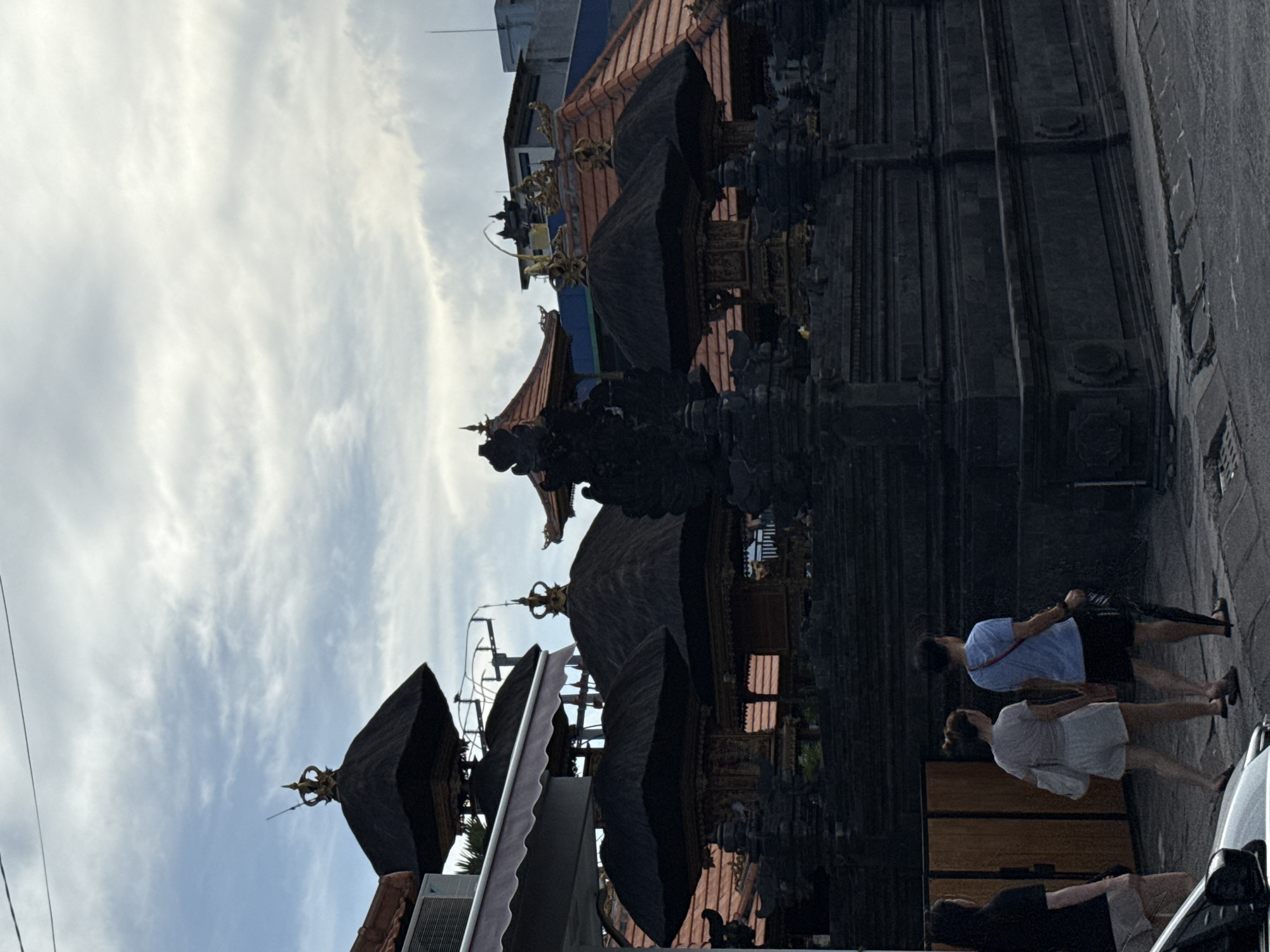 The image shows a street view in North Kuta, Indonesia, featuring a traditional Balinese building with multiple tiers and ornate decorations. The building's dark roof contrasts with the cloudy sky in the background. Several people are walking on the sidewalk next to the building. A portion of a car is visible in the foreground.