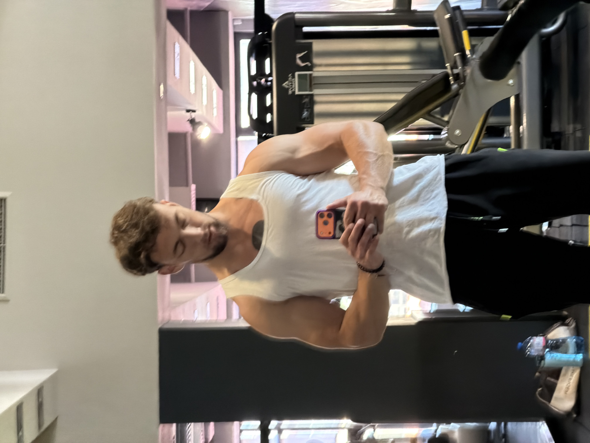 A man in a white tank top and black sweatpants stands in a gym, holding a phone in his hands. He is looking down at the phone. The gym appears to be well-lit with modern equipment. The man has short, curly brown hair and a tattoo on his chest. In the background, there are several pieces of gym equipment and a window. On the floor to the left of the man, there is a gym bag and two water bottles. The scene is consistent with a gym in Tsarevo, Bulgaria.