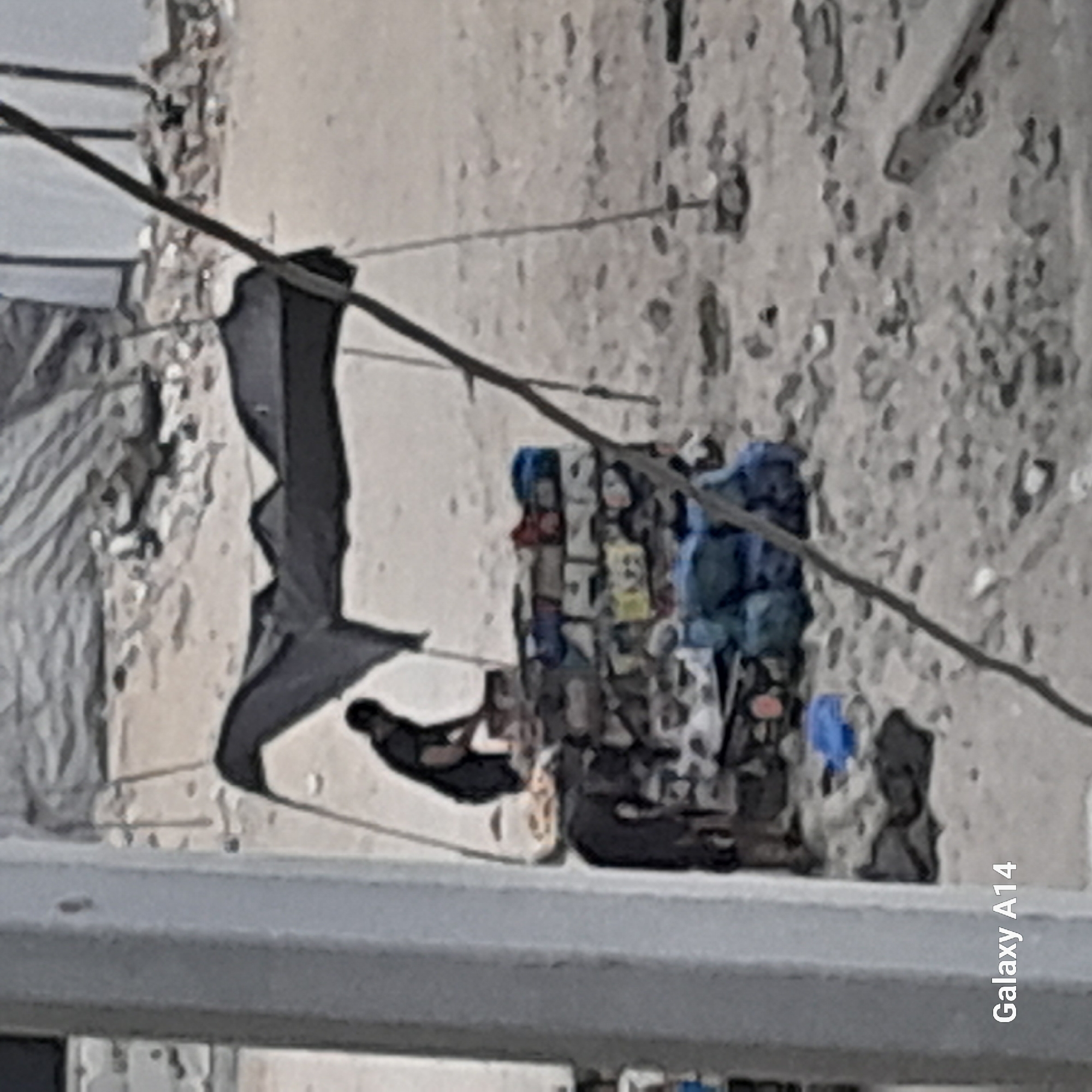 A person, silhouetted in dark clothing, is bent over a makeshift stall covered by a dark tarp supported by poles. The stall appears to be constructed from stacked bricks and is laden with various items, including bags and possibly clothing. Another dark-clothed figure is partially visible on the left, also appearing to be interacting with the stall. The ground is rough and sandy, with scattered debris. The scene suggests an outdoor market or roadside stall. The image contains a watermark indicating "Galaxy A14". The background shows more debris and the blurred outline of structures. The location is identified as Khan Yunis, Palestinian Territories.