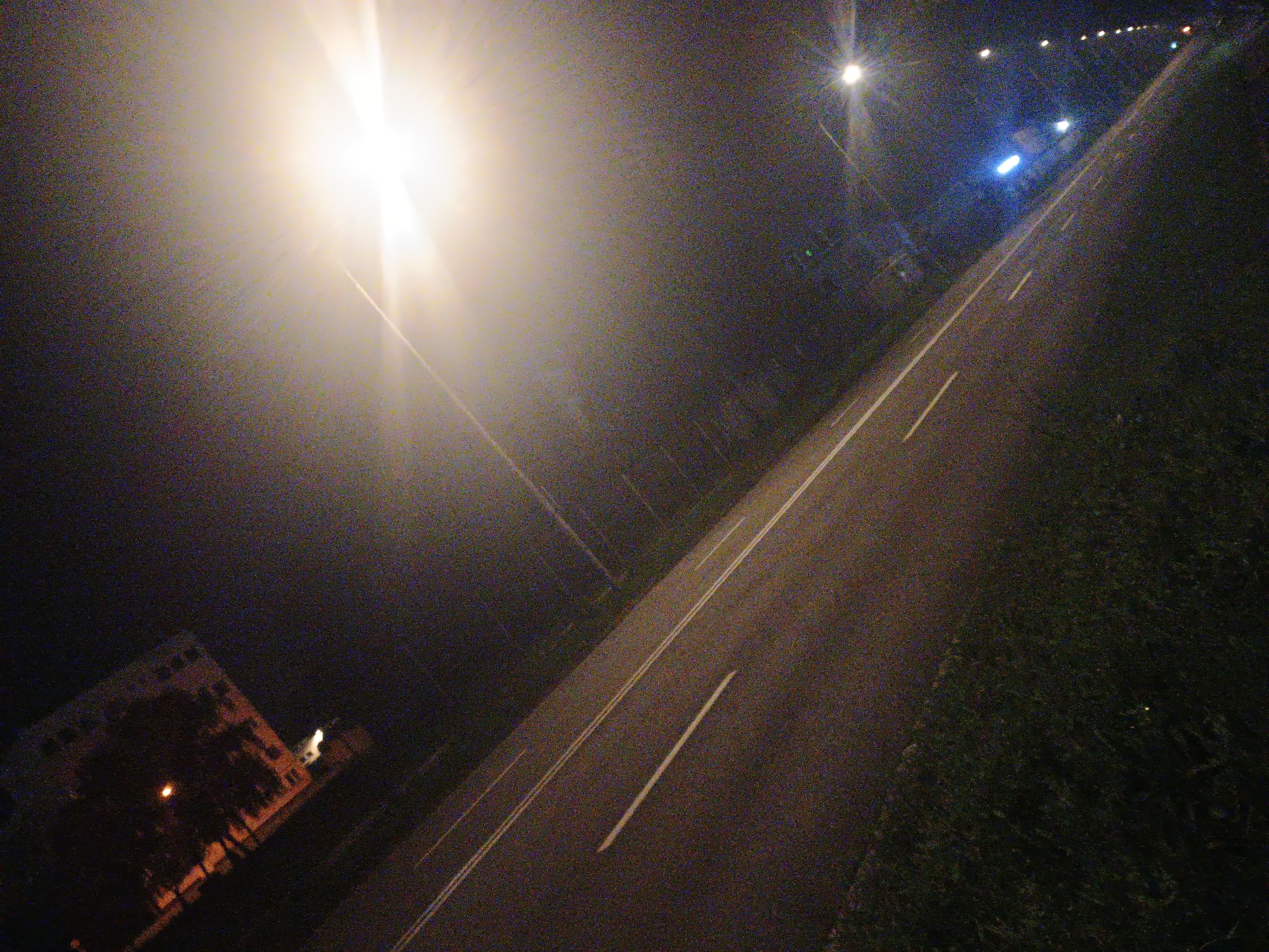 This nighttime image captures a street scene in Kroměříž, Czechia, from an angled perspective. A paved road, marked with white dashed and solid lines, stretches diagonally across the frame from bottom-left to top-right. To the right of the road, dark grass or dense foliage is visible. On the lower-left, a building, possibly an apartment complex, with illuminated windows, is partially obscured by trees.
The scene is primarily lit by artificial light sources. A powerful, brightly glowing streetlight on the mid-left side creates significant lens flare and atmospheric haze, diffusing light across a large portion of the upper-left area. Further down the road, additional streetlights appear as distinct bright points, with one prominent blue light source visible in the distance.
The time of day is night, characterized by low light conditions and noticeable image noise, consistent with night photography. There is no visible activity or event taking place; the street appears deserted without any people or vehicles. No text is visible within the image. The overall atmosphere is quiet and still, enhanced by the artificial illumination against the dark sky.