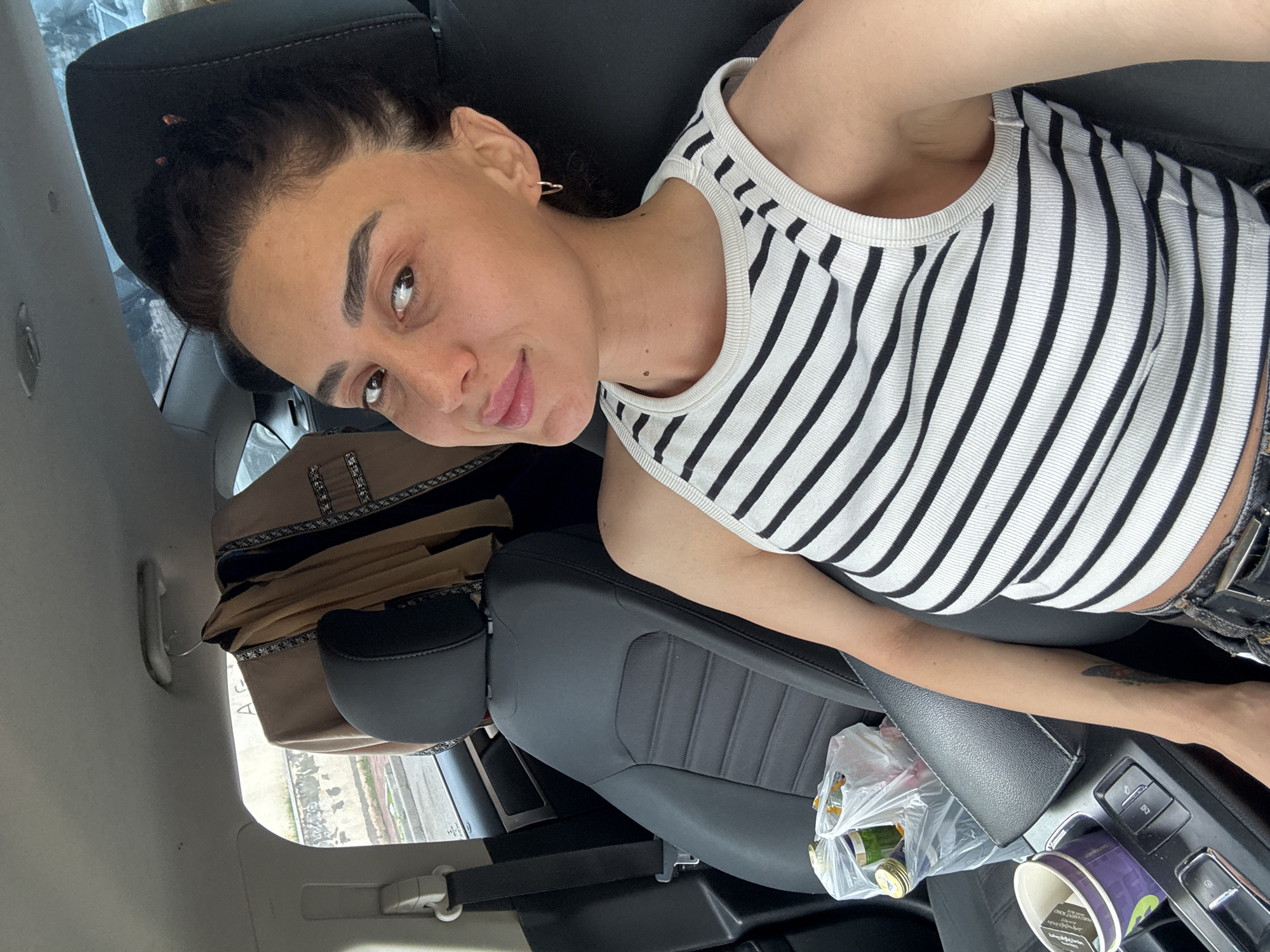 A woman with dark brown hair pulled back in a bun and dark brown eyes is sitting in the passenger seat of a car. She is wearing a black and white striped tank top and dark denim jeans. Her left arm rests on her lap. Next to her is a plastic bag containing bottles and other items, and a purple cup. A beige jacket is hanging on a hanger from the car's ceiling. The car appears to be parked on a street with a building and trees visible through the window. The setting is consistent with Tbilisi, Georgia.
