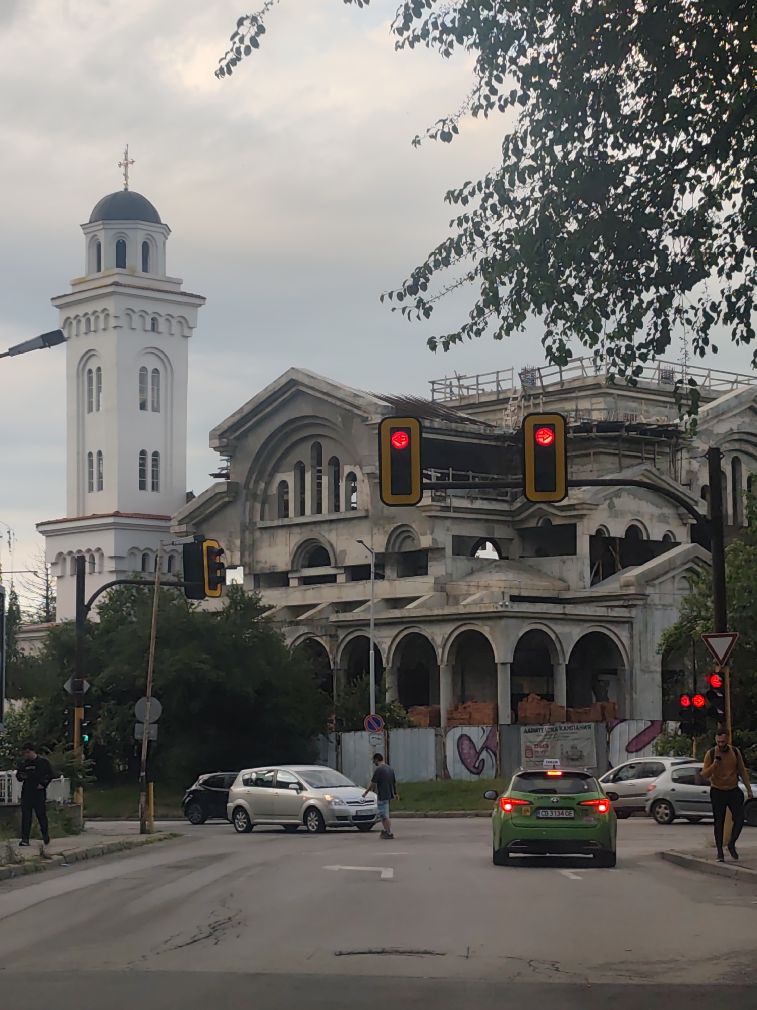 This image depicts a bustling street intersection in Sofia, Bulgaria, under an overcast sky, suggesting either early morning or late afternoon.
In the background, two prominent structures dominate the scene. On the left stands a elegant white church tower, topped with a dark dome and a golden cross. Adjacent to it, a much larger building is under construction or renovation. This structure features multiple arched windows and entrances, exposed concrete, and scaffolding on its upper levels, indicating ongoing work.
At street level, the intersection shows typical urban activity. Traffic lights facing the camera are red, while a green light is visible further left. Several cars are stopped, including a light silver minivan in the foreground, a dark hatchback, and a bright green car displaying the Bulgarian license plate "CB 3134 OE" and what appears to be taxi branding. Its brake lights are illuminated.
Pedestrians are also present. On the left sidewalk, a man in dark clothing is looking at his phone. Another man in a blue shirt and shorts appears to be crossing the street near the silver minivan. On the right sidewalk, a man in a yellow long-sleeved shirt walks while also looking at his phone. A red triangular yield sign is visible on the right. Overhanging tree branches add greenery to the upper part of the image. Visible on a construction fence near the unfinished building is Bulgarian text, partially reading "АДАИТЕЛСКА КАМПАНИЯ" (Advertising Campaign). The scene captures a dynamic urban environment blending historical architecture with ongoing development and daily life.