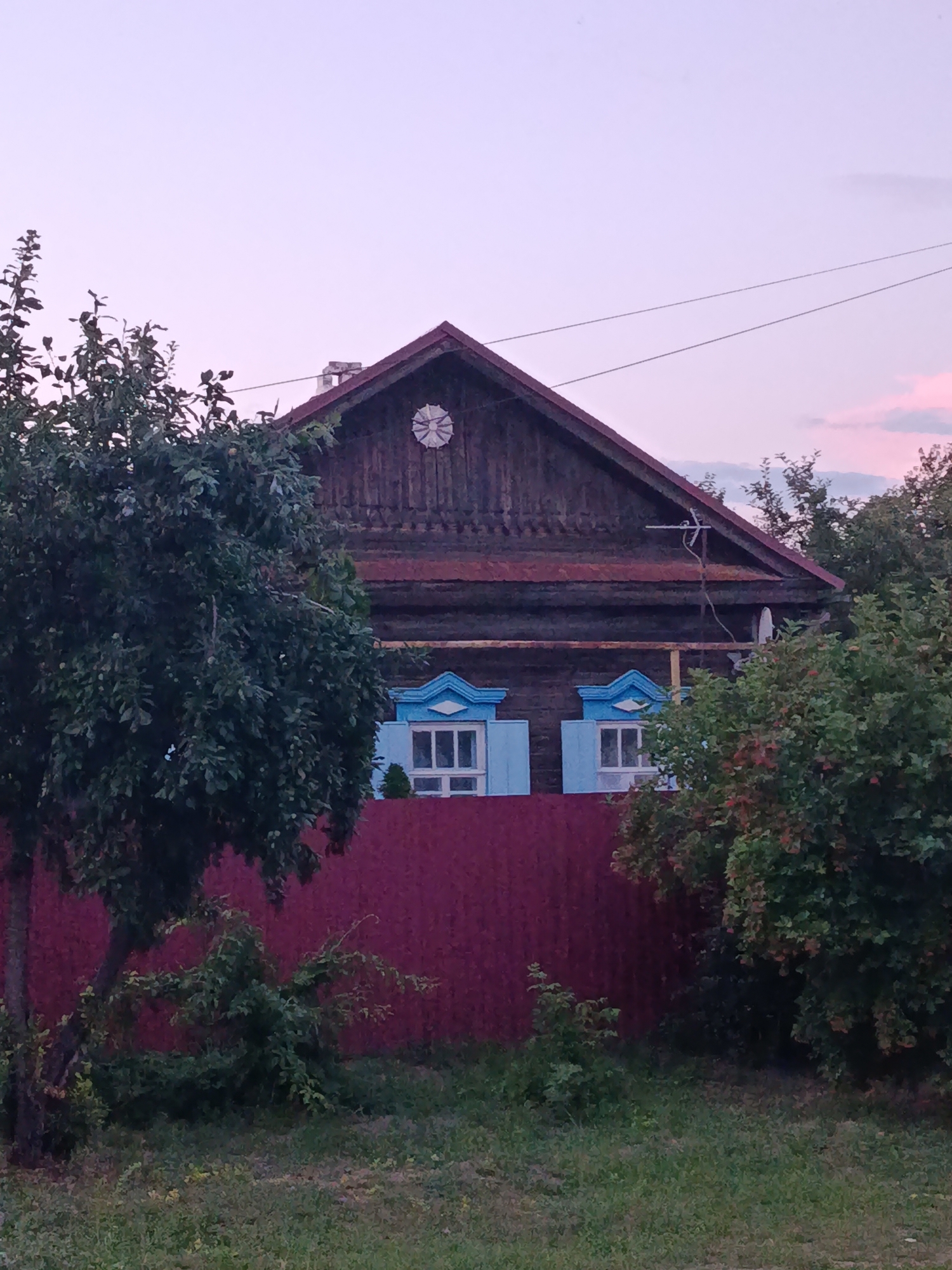 The image shows a wooden house with two windows on the first floor, each with blue shutters. The house has a pitched roof and a chimney. In front of the house is a red fence, and to the left of the house is a tree with green leaves. The sky is a pale pink and purple color, indicating it is either sunrise or sunset. The house appears to be in a rural or suburban setting, possibly in Saratov, Russia, given the architectural style which is common in Russian villages. The overall scene is serene and peaceful, bathed in the soft light of dusk or dawn. There are no people visible in the image. The environment is vegetated with grass and trees.