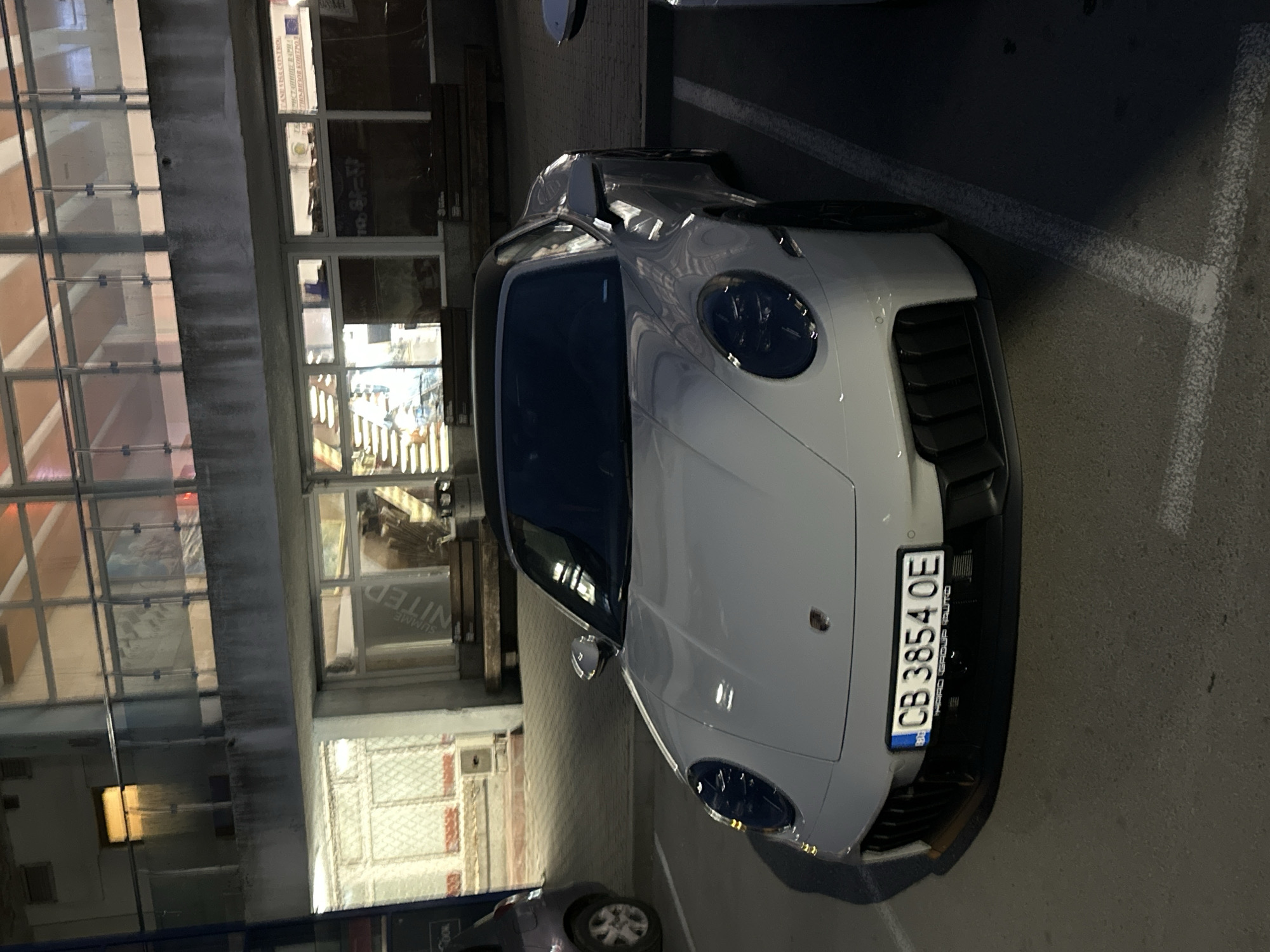 The image shows a white Porsche parked indoors on a concrete surface. The car's license plate is visible and reads "CB 3854 OE". The vehicle is situated near a building, with visible windows reflecting light. There's another car parked nearby in the background. The location may be in Varna, Bulgaria, as the license plate suggests a Bulgarian registration.