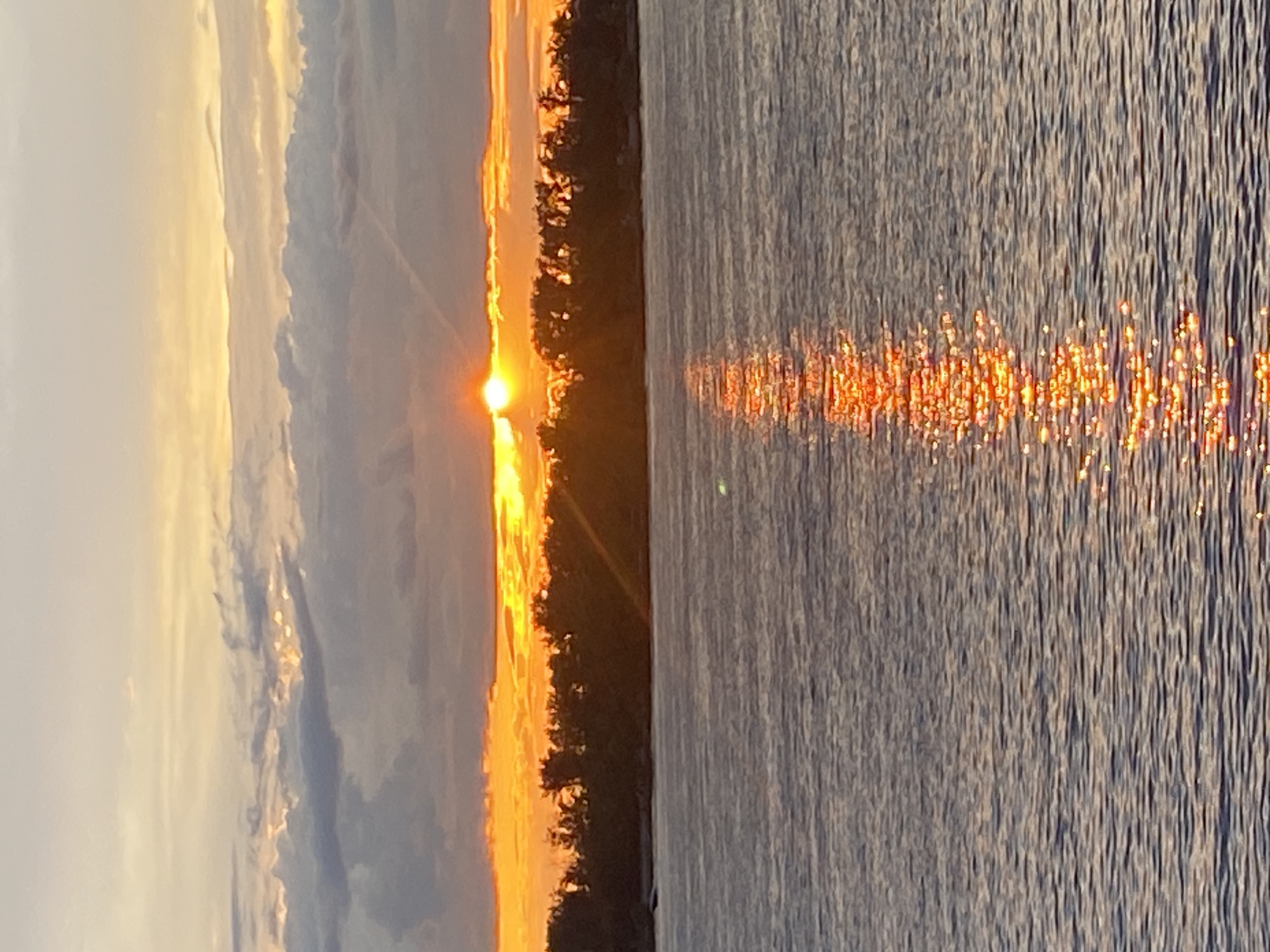 A sunset over the Volga River in Novokuybyshevsk, Russia, creates a vibrant orange glow that reflects on the water's surface. The sky is filled with dramatic clouds, some dark and heavy, others tinged with yellow and orange from the setting sun. A line of trees forms a dark silhouette along the distant bank of the river. The water is calm, with gentle ripples catching the light.