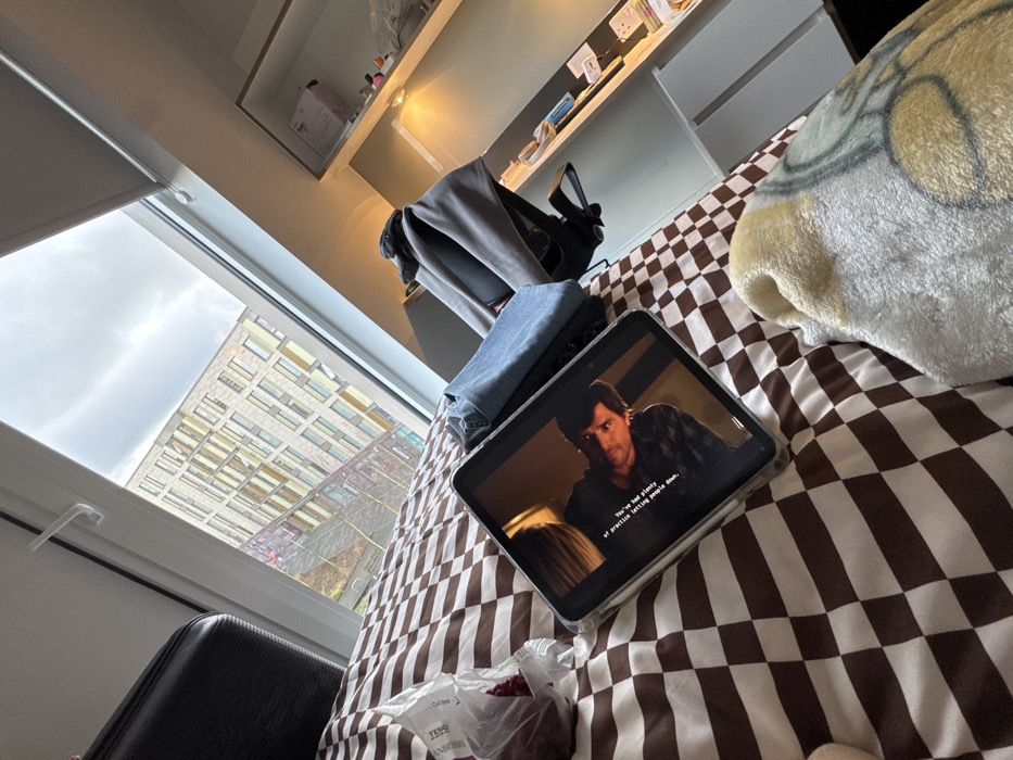 A tablet displays a scene from a movie, resting on a bed with brown and white checkered bedding. A backpack and folded denim sit nearby. A fuzzy, patterned blanket rests beside the tablet. A bag of Tesco cranberries is also on the bed. A window reveals a tall building under an overcast sky. Part of a black chair is visible near the window. Shelving units are attached to the wall.