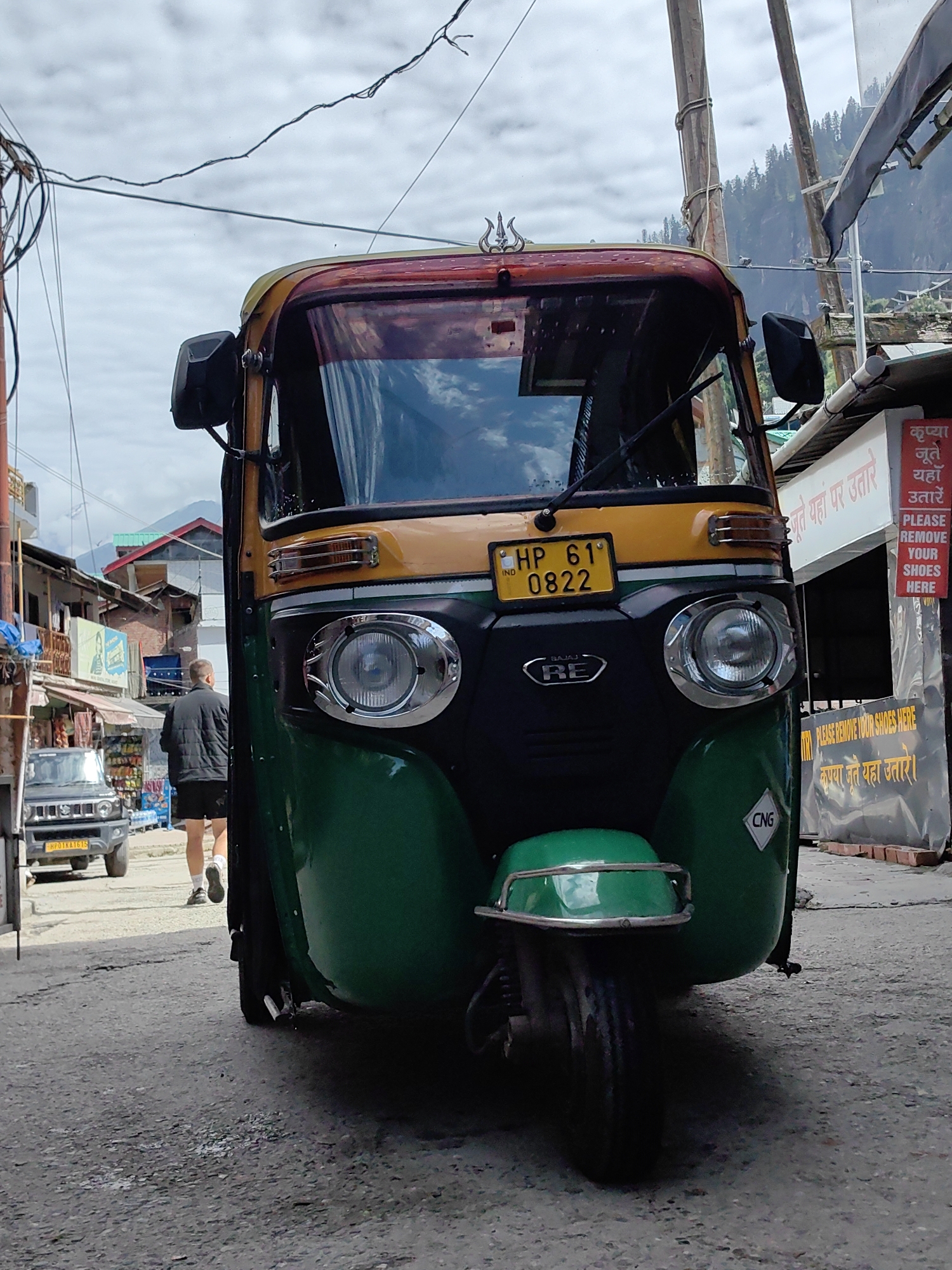 A low-angle, full shot captures a green and yellow auto-rickshaw parked on a paved street in Vashisht, India. The auto-rickshaw dominates the foreground, its front visible with a yellow license plate reading "HP 61 0822". A man wearing a black puffer jacket and shorts walks away from the camera on the left side of the street, past a silver SUV. The background reveals a hillside with sparse trees and buildings, some with red roofs. A sign with Hindi text and "PLEASE REMOVE YOUR SHOES HERE" in English is visible on the right. The sky is overcast.