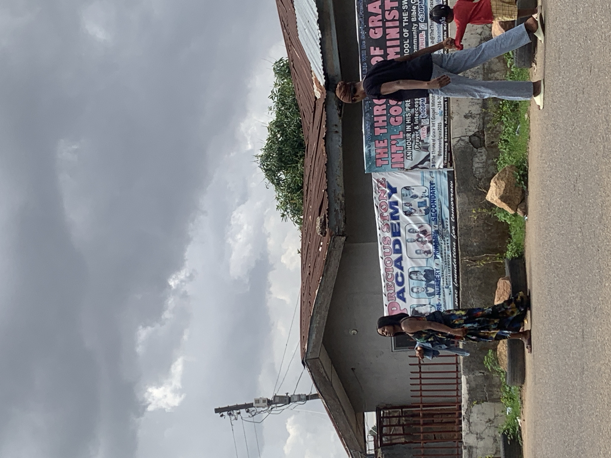 The image shows a street scene with two people in Jos, Nigeria. One person is wearing a long colorful dress, while the other is wearing jeans and a dark shirt. They are standing in front of a building with several signs. The signs are for "Precious Stone Academy" and "The Throne of Grace Int'l Gospel Ministry". The sky is overcast. The text on the sign indicates the ministry is located on Botmang Street. The surrounding environment features weathered buildings and a paved road.