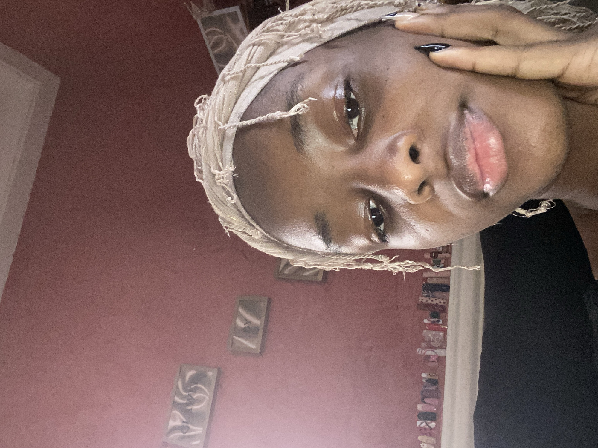 The image is a close-up, indoor, selfie-style photograph of a young Black woman with braids and a light-colored headscarf. She is wearing dark nail polish on her visible fingernail, which is resting on her cheek. The background consists of a textured, pink wall with several framed pictures or artwork hanging on it. Below the wall, a shelf or surface is visible, displaying what appear to be various artificial nails or nail art samples. The lighting suggests it is daytime, possibly with natural light coming from the side. The image quality is moderate, with some graininess. The setting is implied to be a personal space, potentially a room in a home or a salon, located in Jos, Nigeria.