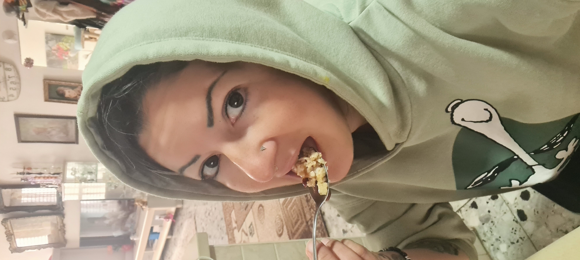 A person with dark hair and brown eyes is eating a forkful of food. They are wearing a light green hoodie with a graphic design on the sleeve. The person has a nose piercing. The background shows a room interior with various decorations, including pictures, mirrors, and a clock. The location is likely Nahariyya, Israel.