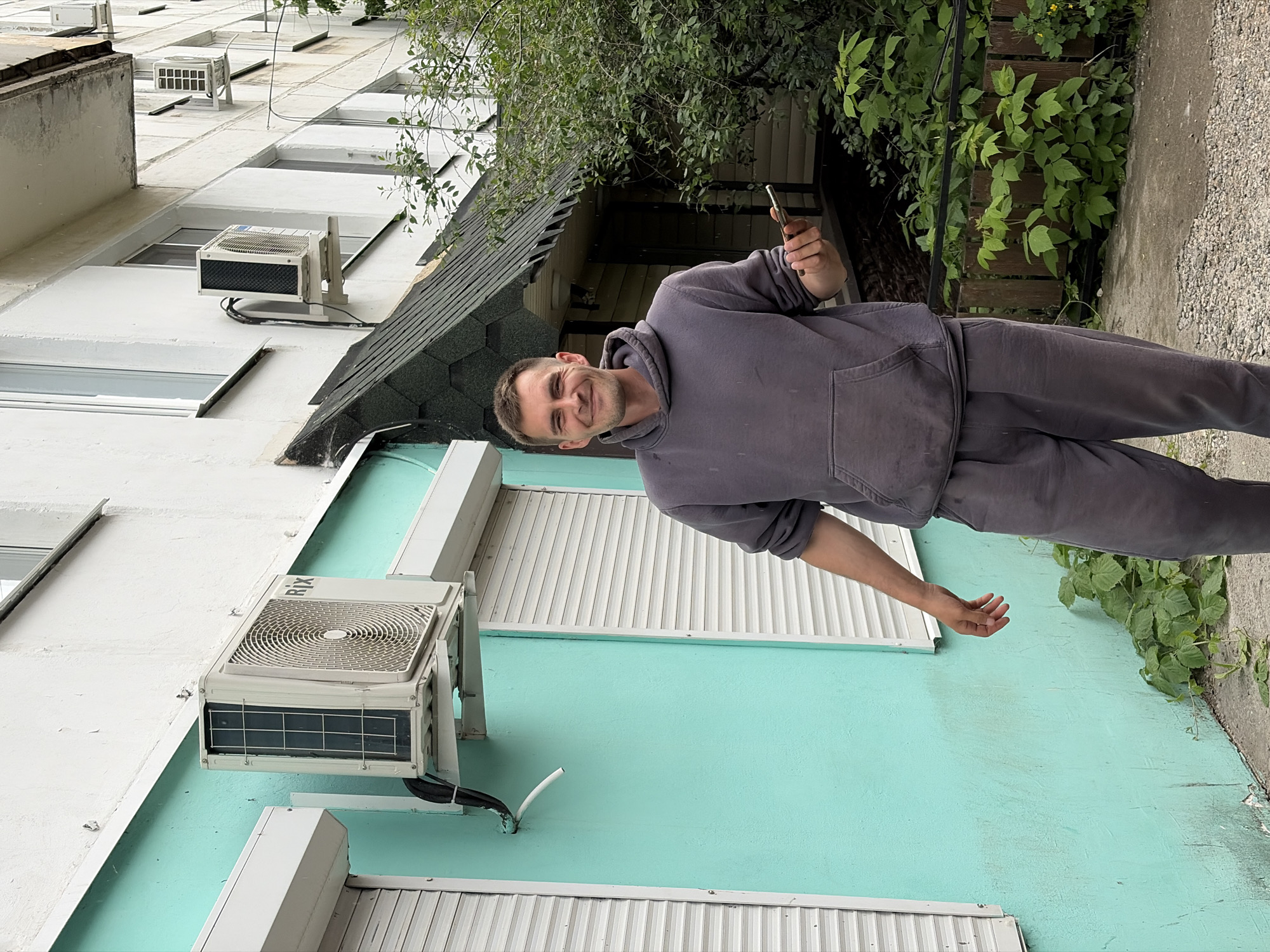 This vertically oriented image (rotated 90 degrees clockwise) features a smiling man standing outdoors against a building. The man has short, dark hair and light stubble, and is wearing a grey hooded sweatshirt and matching sweatpants. He is looking directly at the camera with a broad smile, holding a silver-colored smartphone in his left hand, possibly taking a selfie or video. His right arm is slightly outstretched with an open palm.
The background shows a multi-story building, with its upper levels painted white and the lower section a distinct turquoise color. Several windows are visible, some fitted with white slatted roller shutters. Multiple external air conditioning units are mounted on the building. One unit, prominent on the turquoise section, has the brand name "RIX" visible on its side. A dark, shingled roof structure is partially visible behind the man, suggesting a lower building extension.
To the man's right, there is abundant green foliage, including a leafy tree and ground-level plants, with a wooden fence or barrier partially obscured behind them. The ground surface next to the plants appears to be concrete or gravel. The lighting suggests it is daytime, possibly overcast. The scene depicts a casual moment in an urban or semi-urban environment, characteristic of Chaikovsky, Russia.