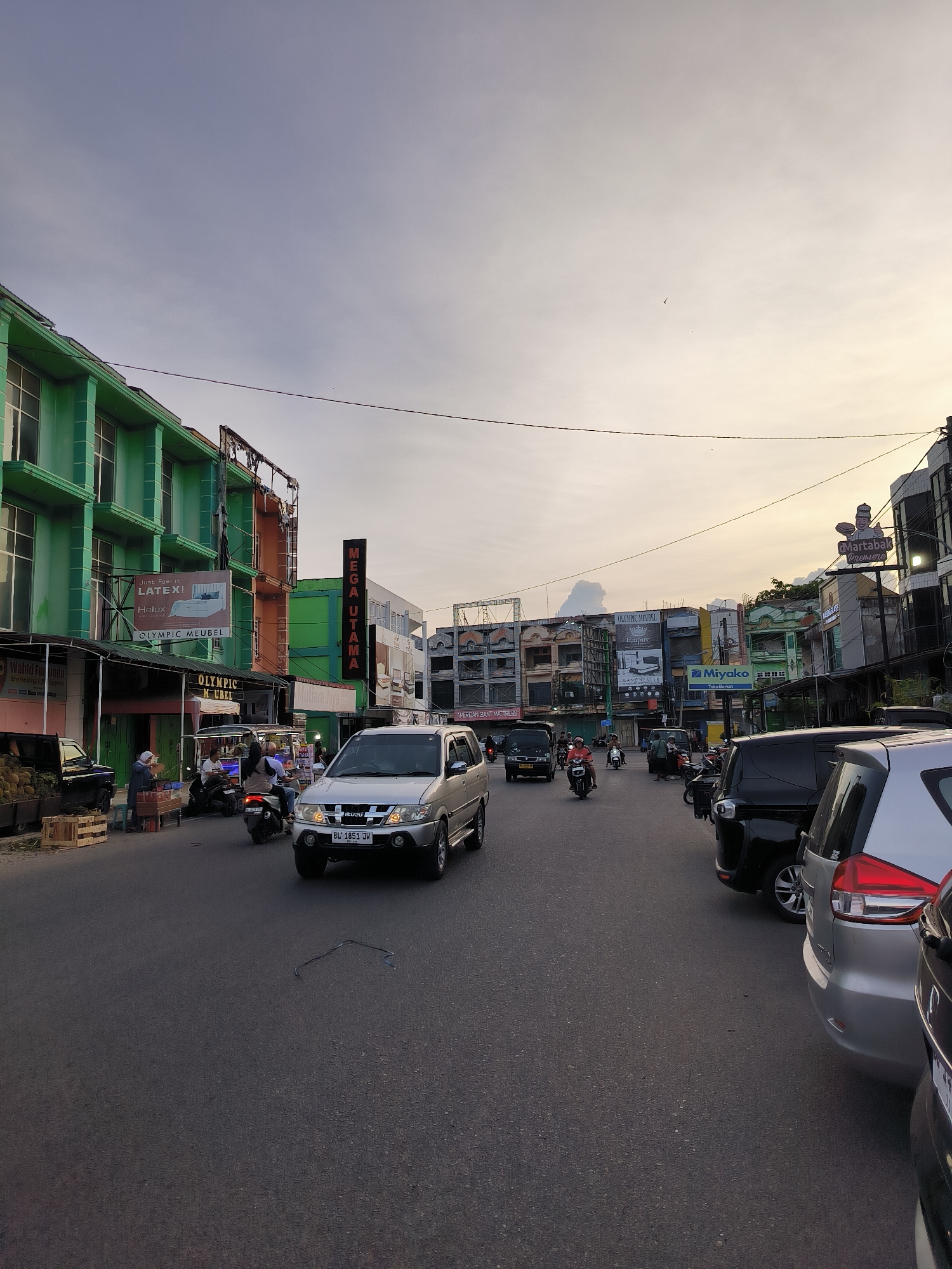 A street scene in Banda Aceh, Indonesia, is depicted at dusk. The street is lined with multi-story buildings exhibiting a variety of architectural styles and colors, including vibrant green and orange facades. Signs for businesses such as "MEGA UTAMA," "Martabak Premiere," "Miyako," and "OLYMPIC MEUBEL" are visible. The sky above is overcast with a soft, warm glow, suggesting the setting sun.
The street is moderately busy with traffic. Several motorcycles are in motion, carrying one or two passengers. A silver Isuzu SUV with license plate BL 1851 JW is prominent in the foreground, heading towards the viewer. Other vehicles, including a black SUV and the rear of a silver car, are parked on the side of the road.
On the left side of the street, stalls are set up, with one displaying a large quantity of durian fruit in wooden crates. A few individuals are seen standing near these stalls, possibly vendors or customers. Overhead, power lines are strung across the scene. The overall atmosphere is that of a commercial area winding down for the day.