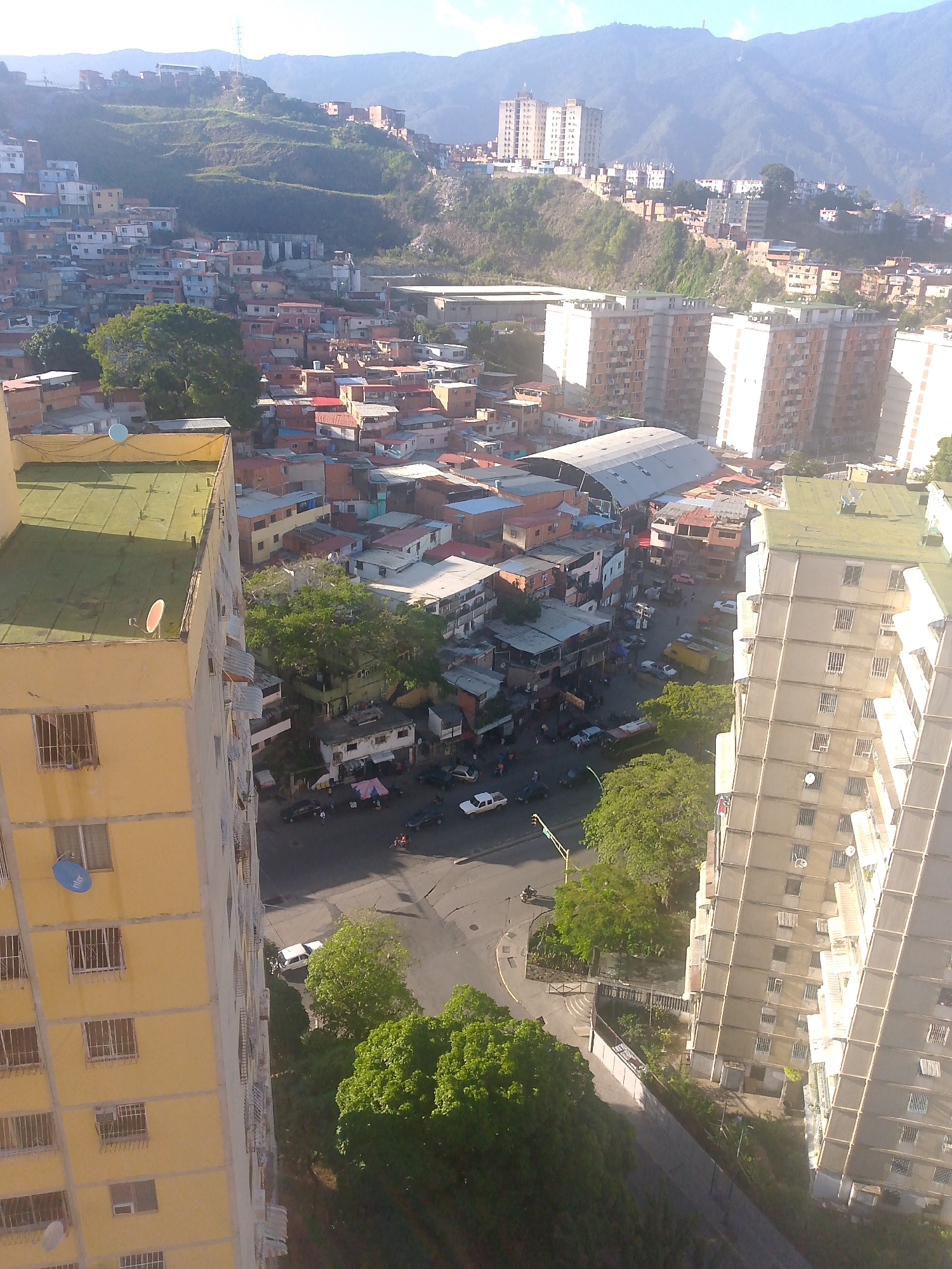 The image provides a high-angle view of a dense urban area in Caracas, Venezuela, characterized by a mix of residential buildings and a hillside favela. In the foreground, a tall, yellow residential building dominates the left side of the frame, its facade featuring numerous windows and balconies. To the right, another tall, light-colored residential building is visible, with rows of windows.
The middle ground reveals a sprawling neighborhood of smaller, brightly colored houses with red-tiled roofs, characteristic of the hillside settlements in Caracas, likely part of the Petare district, known for its extensive favelas built on steep terrain. A large, covered structure, possibly a market or community center, is situated amidst these smaller homes. Several vehicles, including cars, motorcycles, and a yellow truck, are present on the streets and in parking areas, indicating active urban life. Green trees are scattered throughout the scene, providing patches of foliage amidst the concrete and brick structures.
In the background, more residential buildings climb the steep hillsides, leading up to a mountainous landscape that forms the backdrop of the city. The sky is clear, with the sun casting shadows and illuminating the urban sprawl. The overall impression is one of a vibrant but somewhat chaotic urban environment, with a clear contrast between the modern high-rise apartments and the informal settlements on the hills. There are no clearly identifiable individuals in the image.