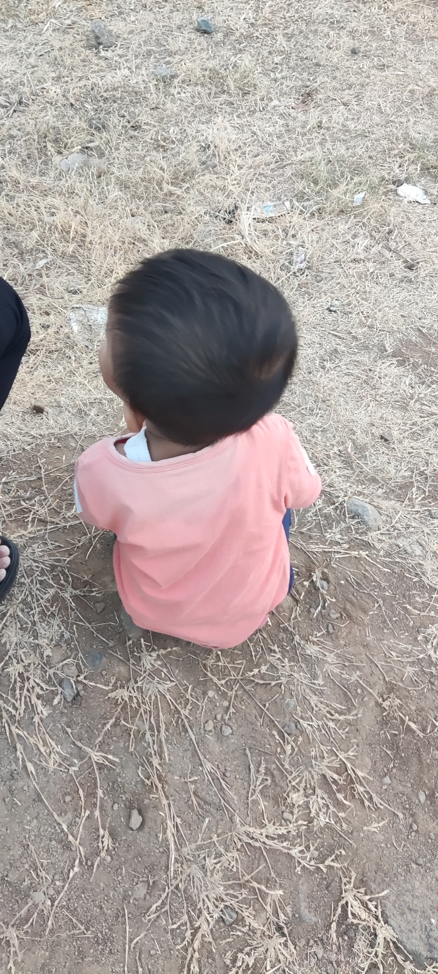 A child wearing a peach-colored shirt and dark pants sits facing away from the camera in a dry, grassy area. The child's hair is dark and appears to be short. The ground is covered in dry grass, dirt, and small rocks. To the left of the child, a black sandal is visible. In the background, there are some scattered pieces of trash, including a white plastic wrapper. The overall environment suggests an outdoor, possibly rural or undeveloped, location. The location is described as Pakusamben, Indonesia.