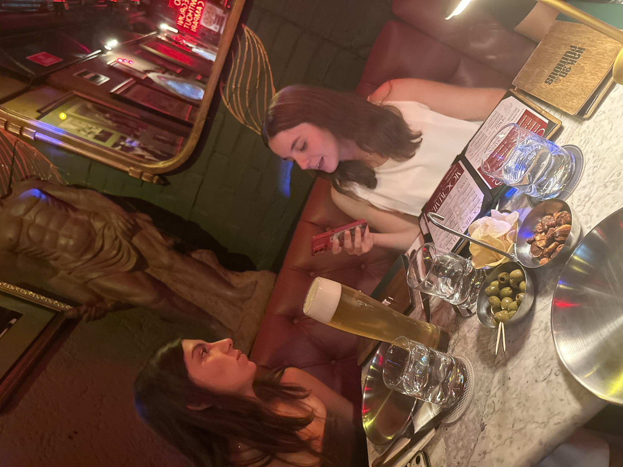 Two women are seated at a table. The woman on the right is wearing a white shirt and holding a red cell phone. The woman on the left is looking at her. The table has a variety of items on it, including a large beer, glasses, bowls of chips, nuts, and olives, a menu, and a "Hak-kan Furious" item. The environment includes an elaborate mirror on the wall with what appears to be artwork underneath it. The restaurant is in the city of Puebla, Mexico.