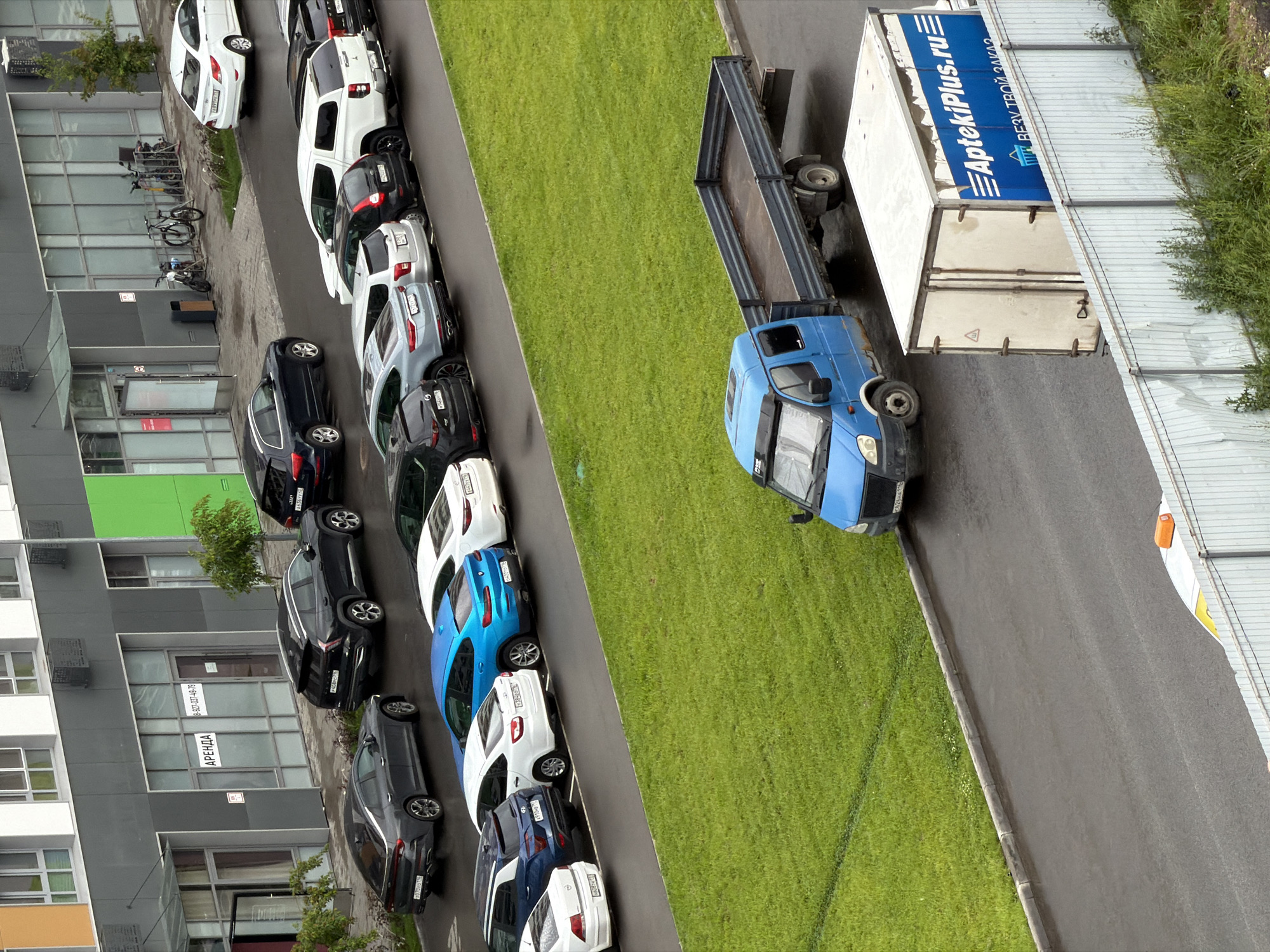 The image shows a street scene with several cars parked along the side of a road next to a grassy area and a modern building. A blue truck with a white cargo container is parked on the road, partially blocking the view of the parked cars. The cargo container has blue and white lettering that reads "AptekiPlus.ru" and "ВЕЗУ ТВОЙ ЗАКАЗ". The building appears to be an apartment complex or office building with large windows and balconies. There are also several bicycles parked near the building. The scene appears to be set in an urban or suburban environment. The time of day and weather are not explicitly clear, but the lighting suggests it could be daytime. There are no people visible in the image.