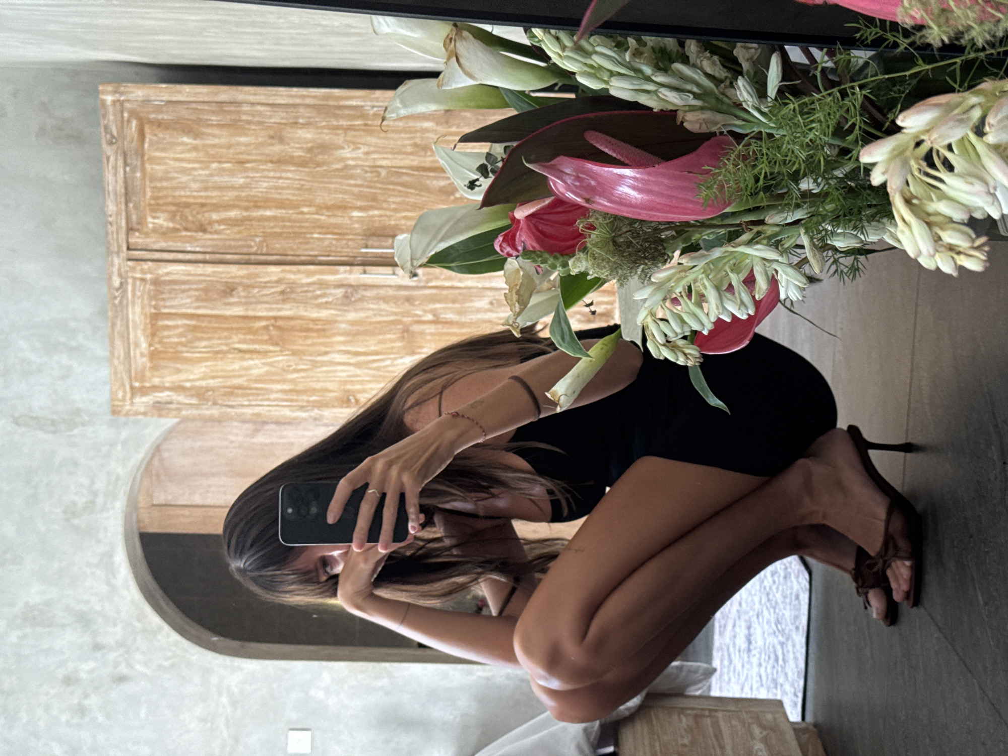 A woman kneels in front of a mirror, taking a selfie with her phone. She is wearing a black dress and brown high heels. The location appears to be indoors in North Kuta, Indonesia, indicated by the tropical flowers arranged near the mirror and the Balinese-style wooden cabinet in the background. The room has a concrete-style finish.