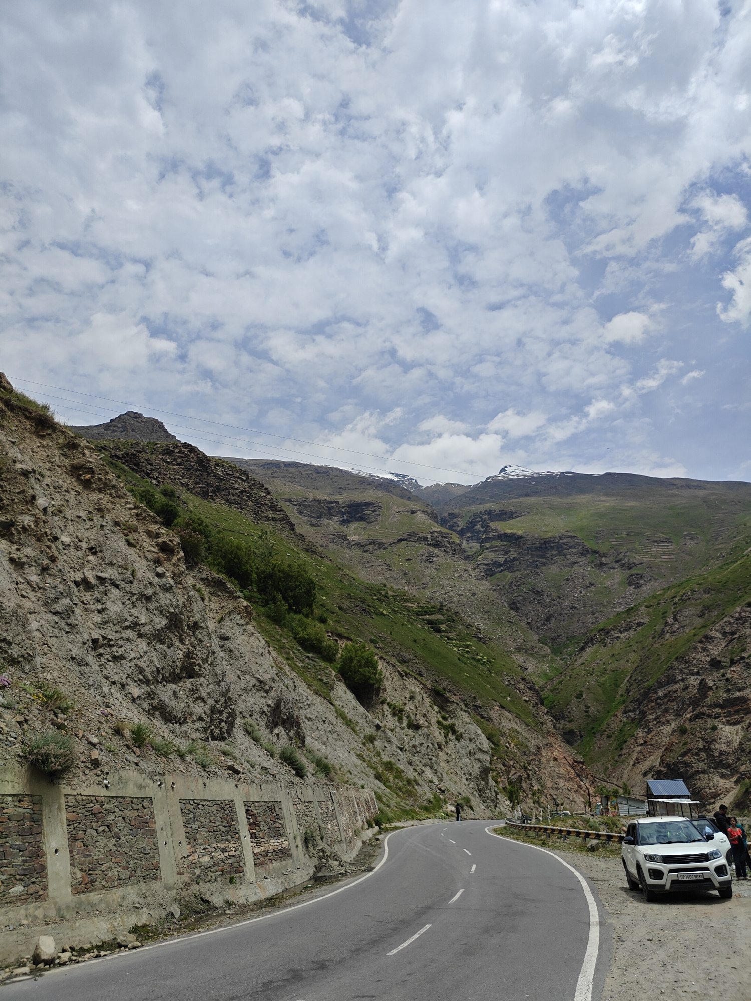 The image captures a scenic mountain road in India, likely in a mountainous region given the terrain and vegetation. The road curves through a valley with steep, rocky slopes on one side and verdant, green mountains in the distance. The sky is partly cloudy with fluffy white clouds against a pale blue backdrop.
On the left side of the road, a retaining wall constructed with stones and concrete supports the rocky embankment, with some sparse vegetation growing from it. Power lines are visible traversing the upper slopes.
On the right side of the road, a white SUV is parked with its doors open, and two individuals are standing near it. Another person can be seen walking on the road further ahead, near a protective barrier along the edge. A small, rustic structure with a blue roof is also visible behind the parked car. The road itself is asphalt with white lane markings. The overall scene suggests a travel or tourist location in a natural, mountainous environment.