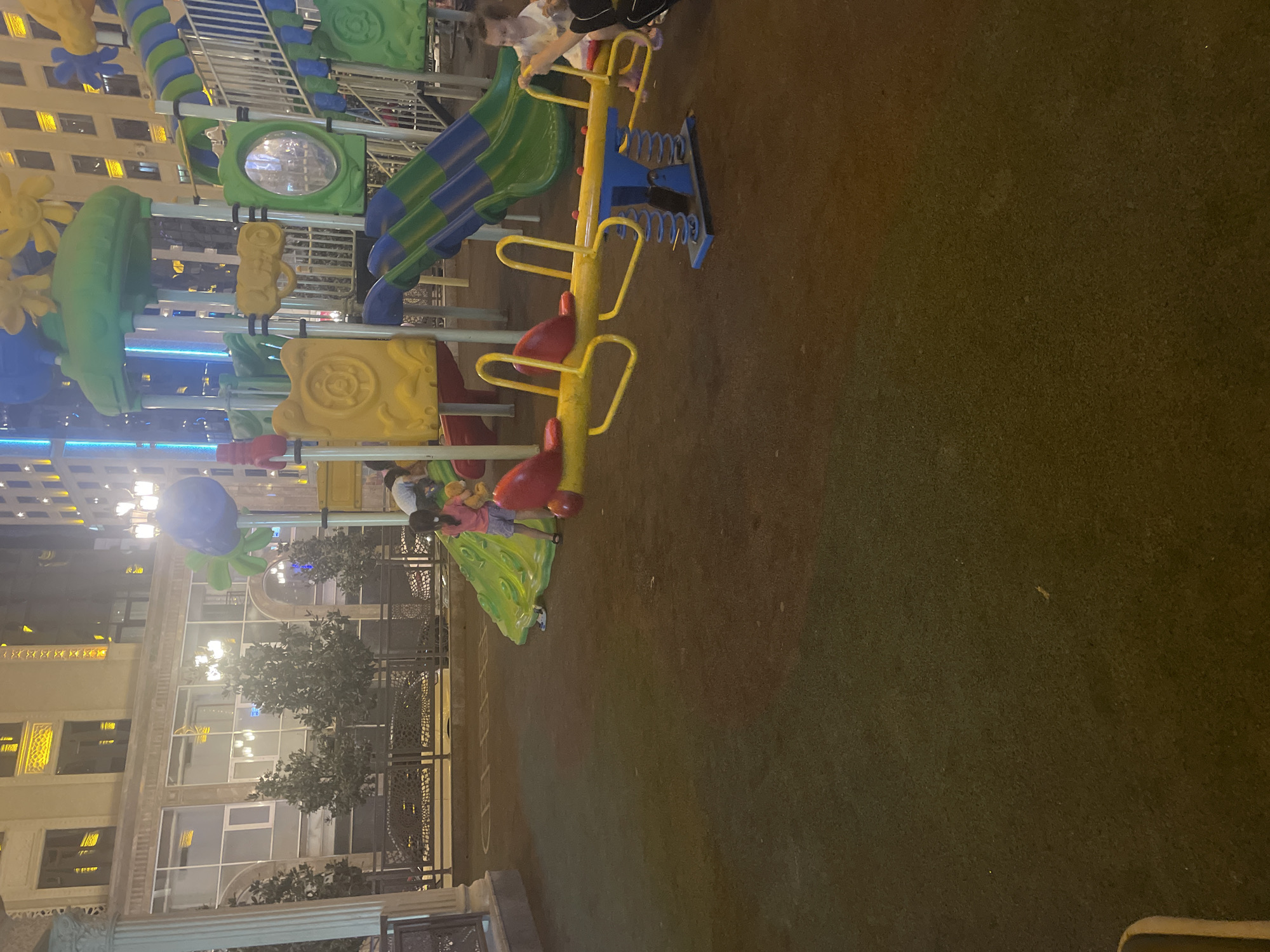 The image depicts a brightly lit playground at night, situated in an urban environment that appears to be the city of Dushanbe, Tajikistan. In the background, a large, modern building with illuminated windows is visible, suggesting a commercial or residential complex. The playground itself is equipped with a colorful, multi-level structure featuring slides, platforms, and climbing elements. A yellow seesaw with blue supports and red seats is prominent in the foreground.
Several individuals, including children, are present. A child in a pink shirt and blue shorts stands near a green slide, facing away from the camera. Another child, wearing a white shirt, is seated on the seesaw, possibly interacting with someone just out of frame. The ground of the playground is covered in a dark, soft material, likely rubber or artificial turf, designed for safety. Trees with dense foliage are planted along a fence in the middle ground, adding a touch of nature to the urban setting. Streetlights and building lights illuminate the scene, creating a vibrant, nocturnal atmosphere. The overall impression is of a well-maintained public space designed for recreation.