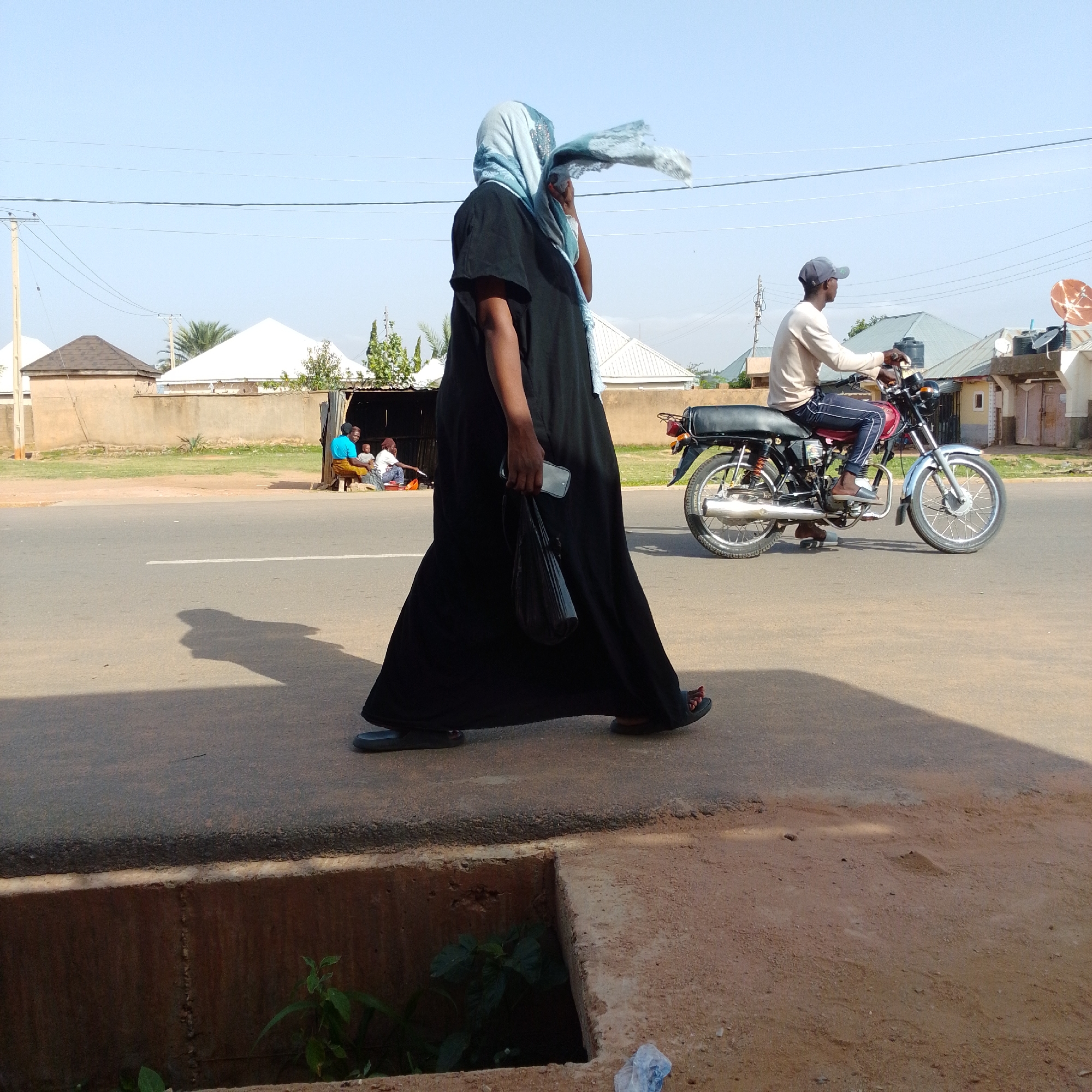 In Gombe, Nigeria, a woman in a black abaya and a light blue hijab walks across a paved road. The wind is blowing her hijab, and she is carrying a black handbag and a mobile phone. To her right, a man wearing a light-colored long-sleeved shirt, dark pants, and a grey baseball cap rides a red motorcycle. Behind the road, buildings with corrugated iron roofs and some concrete structures are visible. A group of people, including two women and a child, are seated in a shaded area next to a roadside structure on the left side of the background. The sky is clear and blue, and the overall environment appears to be an urban or semi-urban setting.