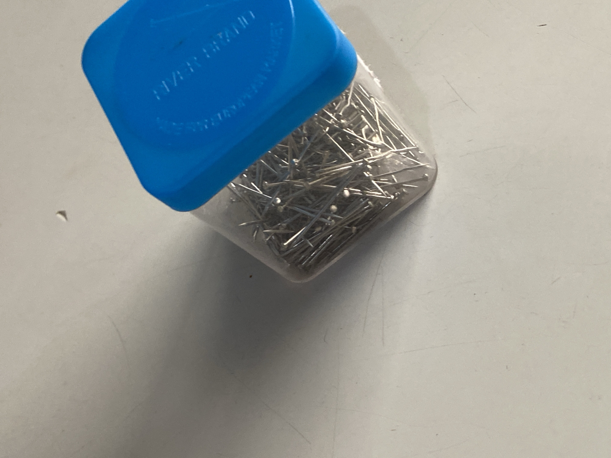 The image shows a clear plastic container filled with numerous silver sewing pins, capped with a bright blue plastic lid. The container is tilted, with the pins spilling slightly over the opening. The lid has embossed text that is partially legible, appearing to say "MADE IN ENGLAND". The container and its contents are resting on a pale, textured surface that appears to be a tabletop or desk. The surface has faint scratches and is unevenly lit, casting a shadow from the container to the left. There are no people or animals visible in the image, and the scene is indoors. The image does not provide any information about the city of Jauro Yinu, Nigeria.