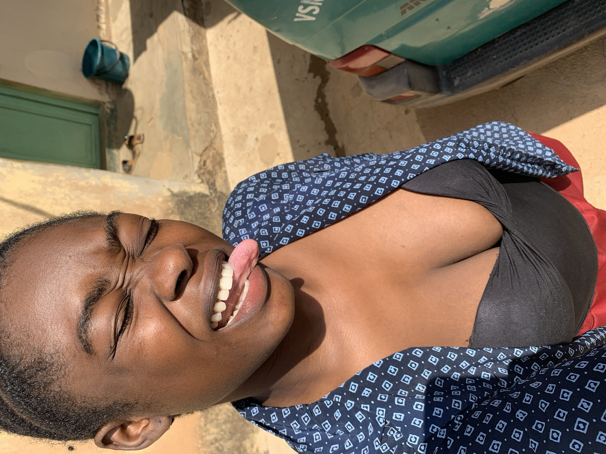 A medium shot captures an African woman outdoors during daylight, presumably in Rafin Par, Nigeria. She is wearing a black top and a blue patterned shirt. Her face is contorted in a grimace, eyes closed, and tongue sticking out. The setting appears to be a street or yard, with a portion of a green vehicle visible at the top, and a green door and a blue bucket in the background. The ground is a mix of concrete and dirt.