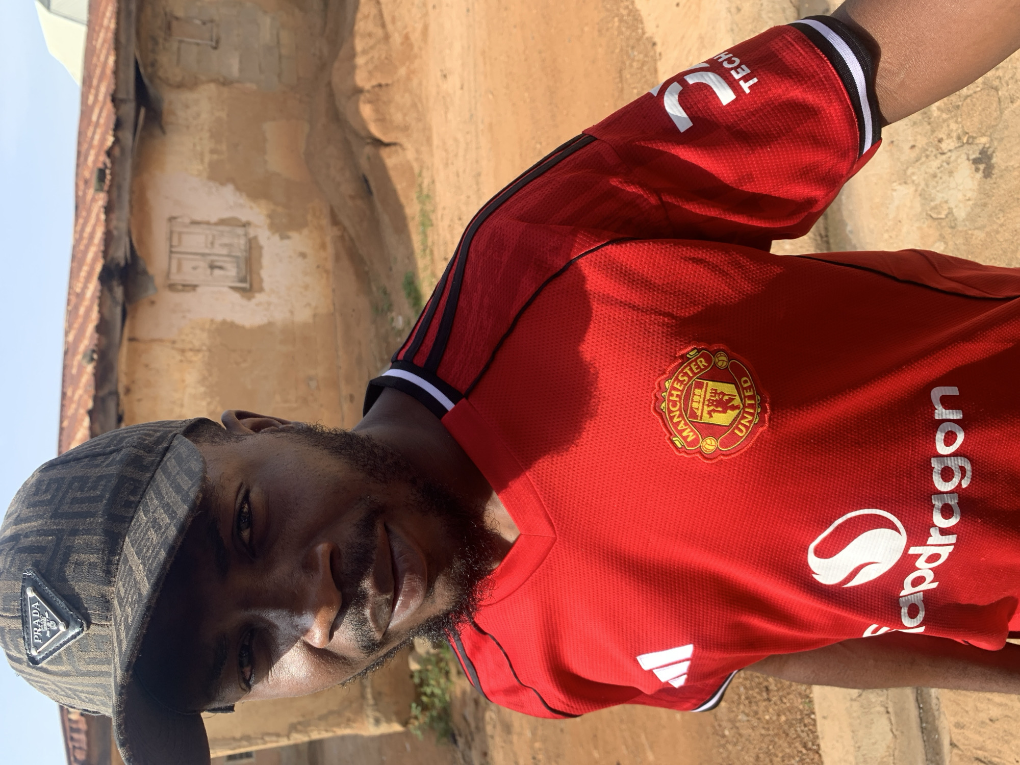 In Rafin Par, Nigeria, a young man with dark skin and a beard stands outdoors, looking towards the camera with a slight smile. He is wearing a red Manchester United jersey with the team's crest and sponsors' logos, including "Snapdragon" and "3 TECH". On his head, he sports a dark baseball cap with the "PRADA" logo. The background consists of a rustic, earthy setting with a weathered, mud-brick house with a corrugated metal roof, suggesting a rural or less developed urban area. The ground is dry and dusty, characteristic of the Nigerian landscape.