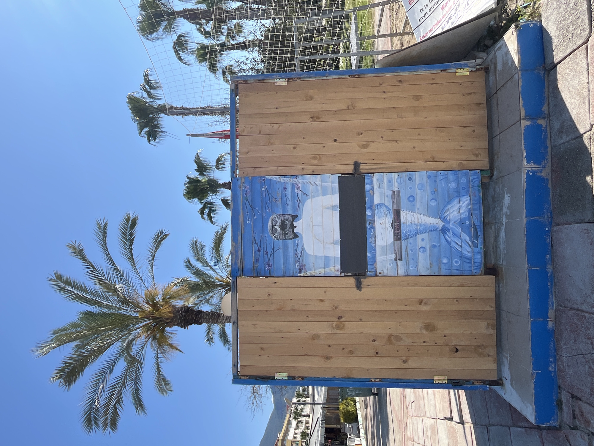 The image shows a wooden structure with a painted image of what appears to be a stylized mermaid or mythical sea creature. The structure is situated outdoors, likely near a beach or seaside area, suggested by the presence of palm trees and the sunny, clear sky. The structure is box-like, with wooden planks and what looks like a blue painted frame. Part of the structure, or perhaps an adjacent surface, is painted with a mural featuring bubbles and the figure. The surroundings include paving stones and other urban elements. It could be a changing room or a small booth in a coastal city, possibly in Alanya, Turkey, as indicated in the prompt. There are no people visibly depicted in the photograph. The image composition has an upward perspective and is tilted.