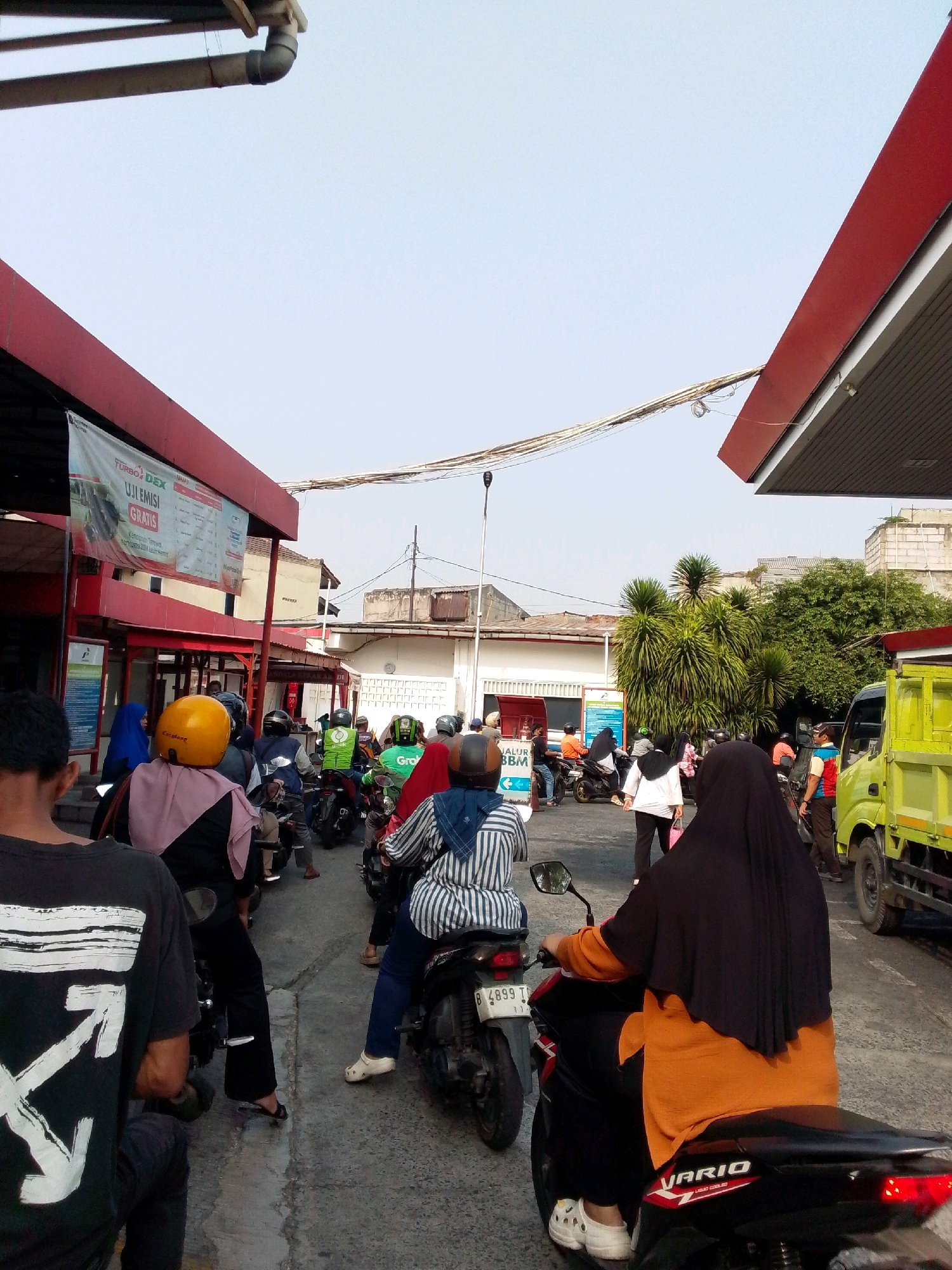 This is a full, vertical shot of a gas station queue in East Jakarta, Indonesia. The scene is dominated by a long line of motorcycles and their riders, all waiting to refuel. The gas station structure, with its red roof and white pillars, frames the left and right sides of the image. In the background, the sky is clear and pale, with a few electrical wires strung overhead.
The foreground is occupied by the backs of several individuals on motorcycles. The motorcycle on the far left has a rider wearing a black t-shirt with a white graphic design. Beside them, a person in a pink headscarf and black pants is seated on a motorcycle. Further into the queue, a rider wears a yellow helmet and a person behind them is wearing a blue headscarf. Another rider sports a green jacket with a prominent "Grab" logo.
The middle ground features more motorcycles and people. Several individuals are wearing hijabs in various colors, including blue, red, and black. A bright yellow truck is visible on the right side of the image, parked at the gas pumps. A gas station attendant in a blue and red uniform stands near the truck.
The environment appears to be a typical urban setting, with buildings and trees visible in the distance. The ground is paved with asphalt, and there are several signs and banners around the gas station, one of which advertises "TURBO DEX" and "UJILEMISI GRATIS". The overall impression is one of a busy, everyday scene at a gas station.