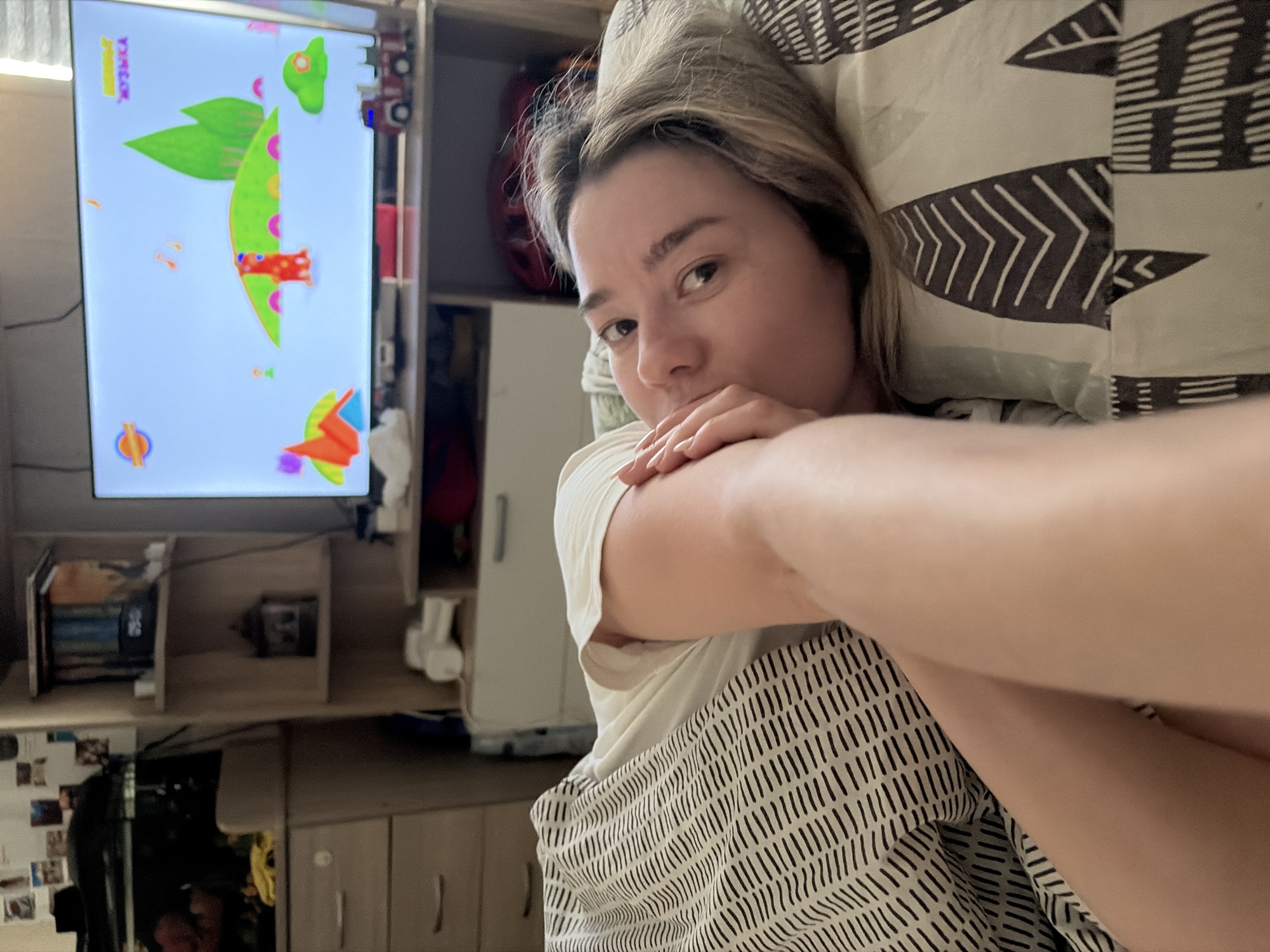 A woman with blond hair is lying in bed with a pillow, appearing to take a selfie. She is wearing a white t-shirt and a striped blanket. The location is in a room with a TV showing an animated program, a bookshelf, and cabinets. Based on the "Музыка" text on the TV screen, the location is likely in a Russian-speaking area. Given the association with Chaikovsky, the city is likely Chaikovsky, Russia.