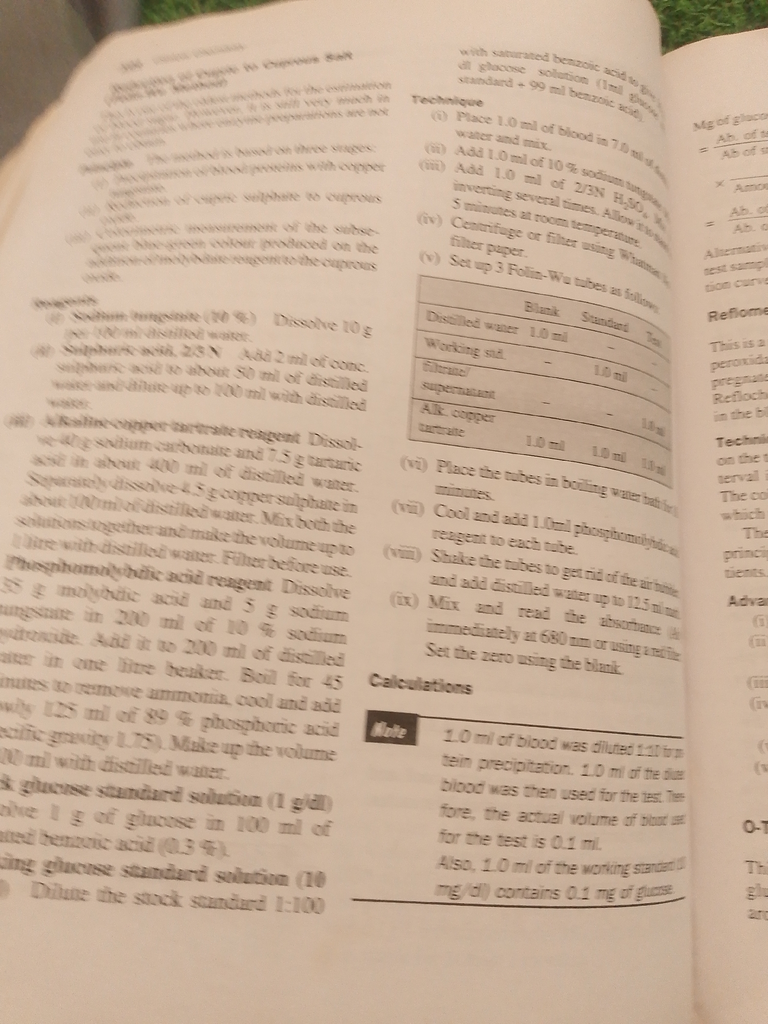 This is a close-up, angled photograph of an open textbook. The book is lying flat on a surface that appears to be a grassy area, suggesting it might be outdoors. The pages are filled with dense text, scientific diagrams, and a table with columns labeled "Blank," "Standard," and "Test." The text appears to be a scientific or medical procedure, likely related to laboratory analysis, as indicated by terms like "reagent," "dissolve," "distilled water," "absorbance," and "glucose standard solution." There is also a section titled "Technique" with numbered steps and a table detailing volumes of different substances. In the upper right corner of the page, there are mathematical formulas and abbreviations like "Mg" and "Ab." A small, rectangular inset box labeled "Note" contains a paragraph of text describing a dilution factor and the actual volume of blood used in a test. The overall impression is that of a practical, instructional manual being used in a potentially informal outdoor setting. The lighting suggests it is daytime.