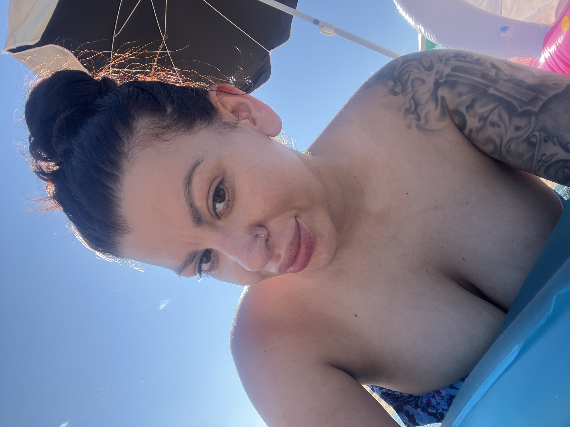 A low-angle, close-up selfie shows a woman in a light blue swimsuit. She has her dark hair pulled up in a bun. Her head is tilted slightly to the right, and she is looking directly at the camera with a slight smile. She has a nose ring and a tattoo on her right arm and shoulder. She is leaning on a bright blue inflatable object, and a white umbrella is visible in the background. The sky is clear and blue. The image appears to have been taken outdoors on a sunny day, possibly near a beach or pool, given the swimsuit and inflatable object. The specific location in Sozopol, Bulgaria, cannot be determined from the image alone.