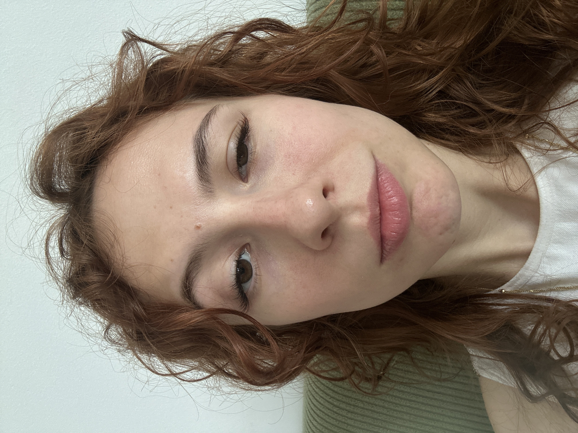 A close-up portrait features a young person, likely female, with reddish-brown, wavy, shoulder-length hair. The subject's head is tilted, giving the impression of looking up towards the camera with a neutral to gentle expression. Their dark brown eyes are open and directed at the viewer, highlighted by prominent eyelashes. The skin appears natural, showing a few small moles or freckles on the forehead and cheek. The lips are a soft pink.
The individual is wearing a white T-shirt, with a thin gold chain necklace visible around the neck. A green, ribbed, knitted material, possibly a sweater or a blanket, is seen along the right side of the frame. The background is a plain, light-colored wall, indicating an indoor setting. The lighting is soft and even, but the specific time of day is not discernible. No activity other than posing for the photograph is depicted, and there is no visible text within the image. The image context is Novi Sad, Serbia.