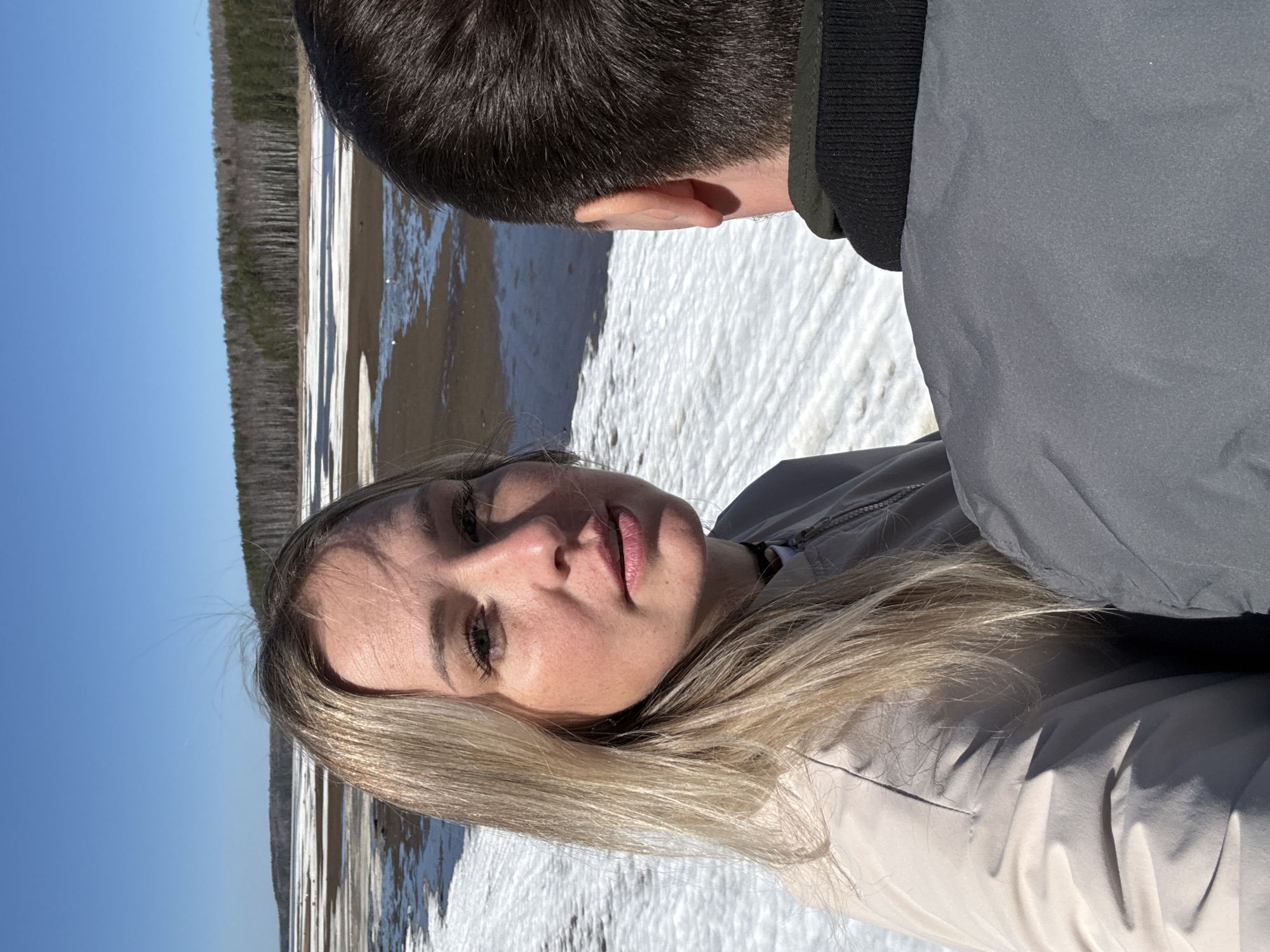 Two people are present in an outdoor environment. A woman with blonde hair is visible in the foreground, wearing a light-colored jacket. The background reveals a landscape with water, snow, and a forest in the distance. The location is identified as Chaykovsky, Russia. The other person is visible from the back of the head.