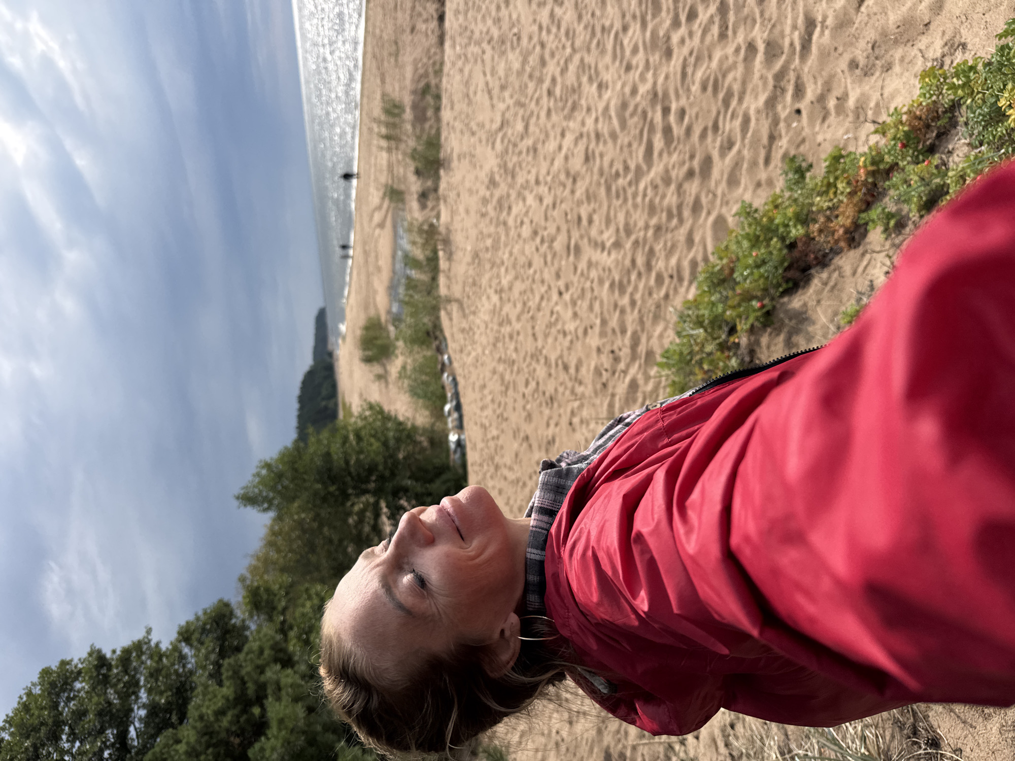 A woman stands on a sandy beach, facing away from the camera and looking towards the water. She is wearing a red hooded jacket over a plaid shirt and has her eyes closed, tilting her head back slightly as if enjoying the sun. The beach is wide and sandy, with sparse vegetation along the edge. In the distance, the sea stretches out to the horizon under a cloudy sky, and a few silhouetted figures can be seen walking along the shoreline. The location appears to be the coast of Zelenaya Roshcha, Russia.