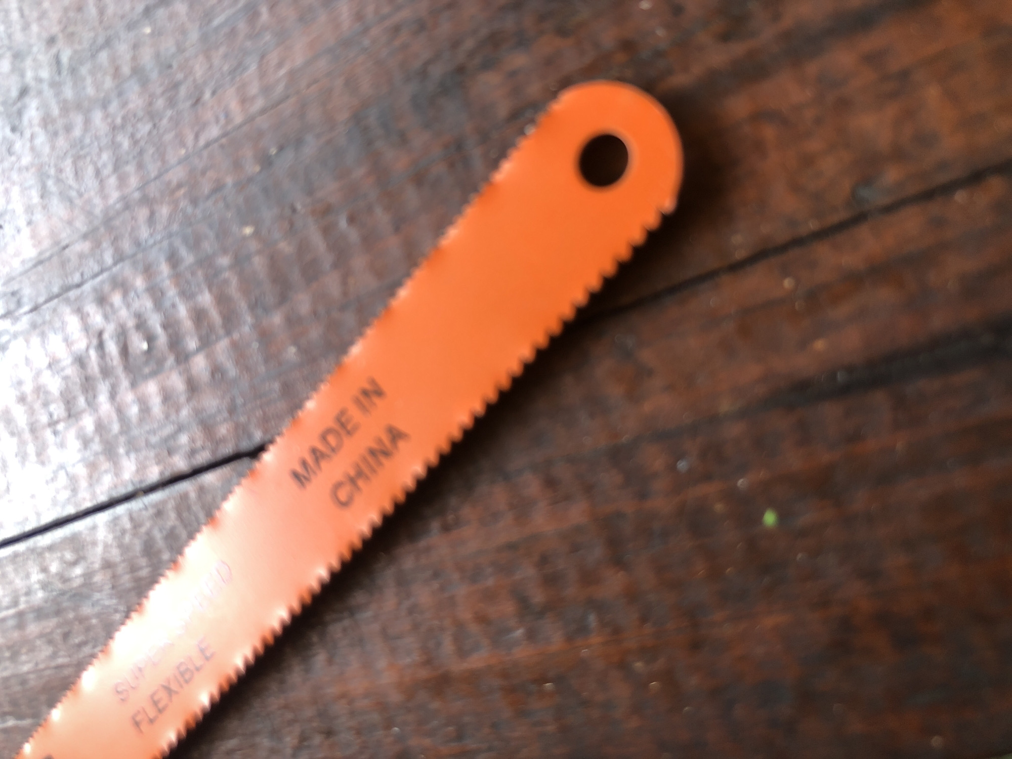 An orange hacksaw blade lies on a dark wooden surface, possibly a table. The blade is angled diagonally across the frame, with the teeth facing upwards and towards the left. Text is visible on the blade, reading "SUPER HARD FLEXIBLE" on the left side and "MADE IN CHINA" on the right. The wooden surface shows grain and has a slightly worn appearance. The scene is lit from above, casting some light and shadow. There is a circular hole at the end of the blade. No people are visible in the image. The location is not specified as Surabaya, Indonesia.