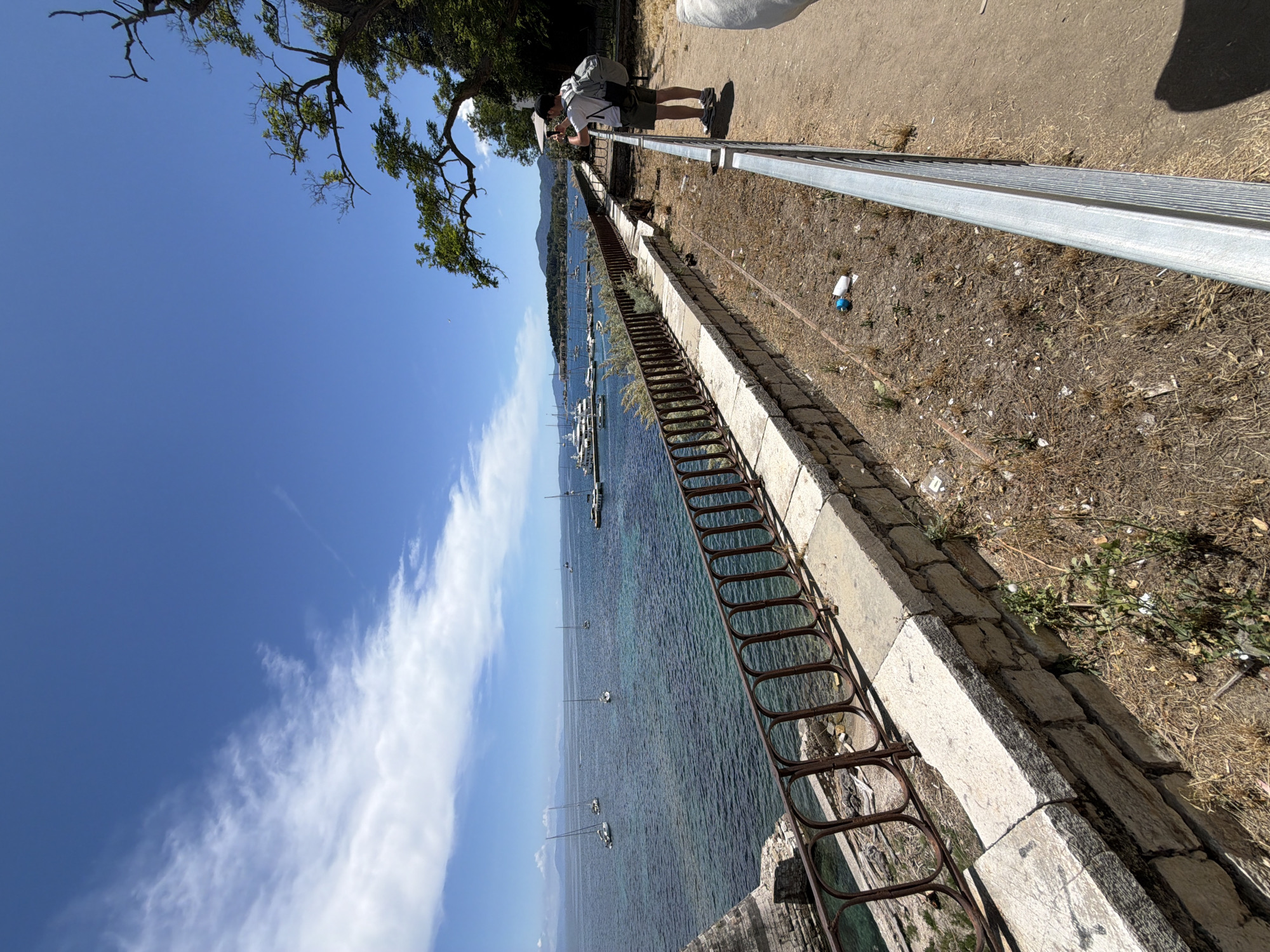 This image, rotated 90 degrees clockwise, depicts a scenic coastal view from an elevated pathway in Corfu, Greece, under a clear blue sky.
In the mid-ground, a person wearing a white t-shirt, dark shorts, a black cap, and a grey backpack is seen standing on a dirt path. They are leaning against a metal railing, appearing to observe the vista. Another partial figure or object, white in color, is barely visible in the upper right.
The path is unpaved, covered with dry earth and sparse vegetation. A discarded white and blue plastic bottle lies on the ground near the railing. The path is separated from the drop to the sea by a two-part barrier: a sturdy, galvanized metal railing forms the top section, and below it, an older, rusty decorative metal railing extends down. A substantial stone retaining wall forms the base of this structure, sloping towards the water.
The sea, a clear blue-turquoise, stretches out, appearing calm and inviting. Numerous boats, including several sailboats and yachts, are scattered across the water, anchored or moored. In the far distance, across the body of water, a range of mountains or hills contributes to the picturesque backdrop, characteristic of the Mediterranean coastline.
The sky is a brilliant blue with a few streaks of wispy white clouds, indicating fair weather. Lush green tree branches partially overhang the scene from the upper left. The strong sunlight and clear shadows suggest the photograph was taken during the middle of the day or early afternoon. The scene conveys a sense of peaceful natural beauty and a pleasant, sunny atmosphere. No visible text is present in the image.