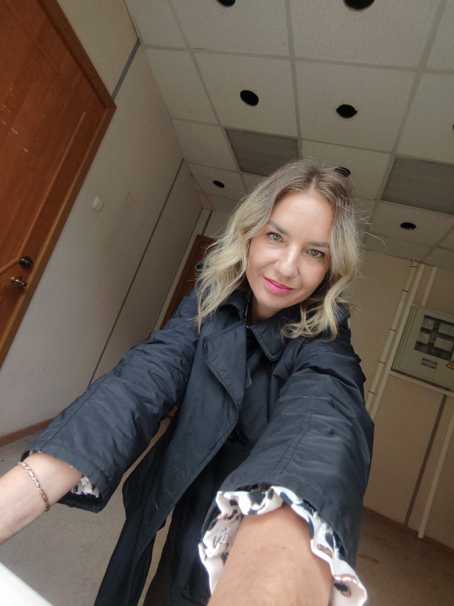 A selfie taken indoors in Petropavlovsk-Kamchatsky, Russia. The subject is a blonde woman wearing a black jacket with a patterned white and black lining visible at the cuffs. She is wearing pink lipstick and a gold bracelet on her left wrist. The background includes a hallway with a drop ceiling with circular light fixtures. A wooden door is visible to the left, and an electrical panel is on the wall to the right. The walls are a light beige color. The floor is gray. The overall lighting is fluorescent.