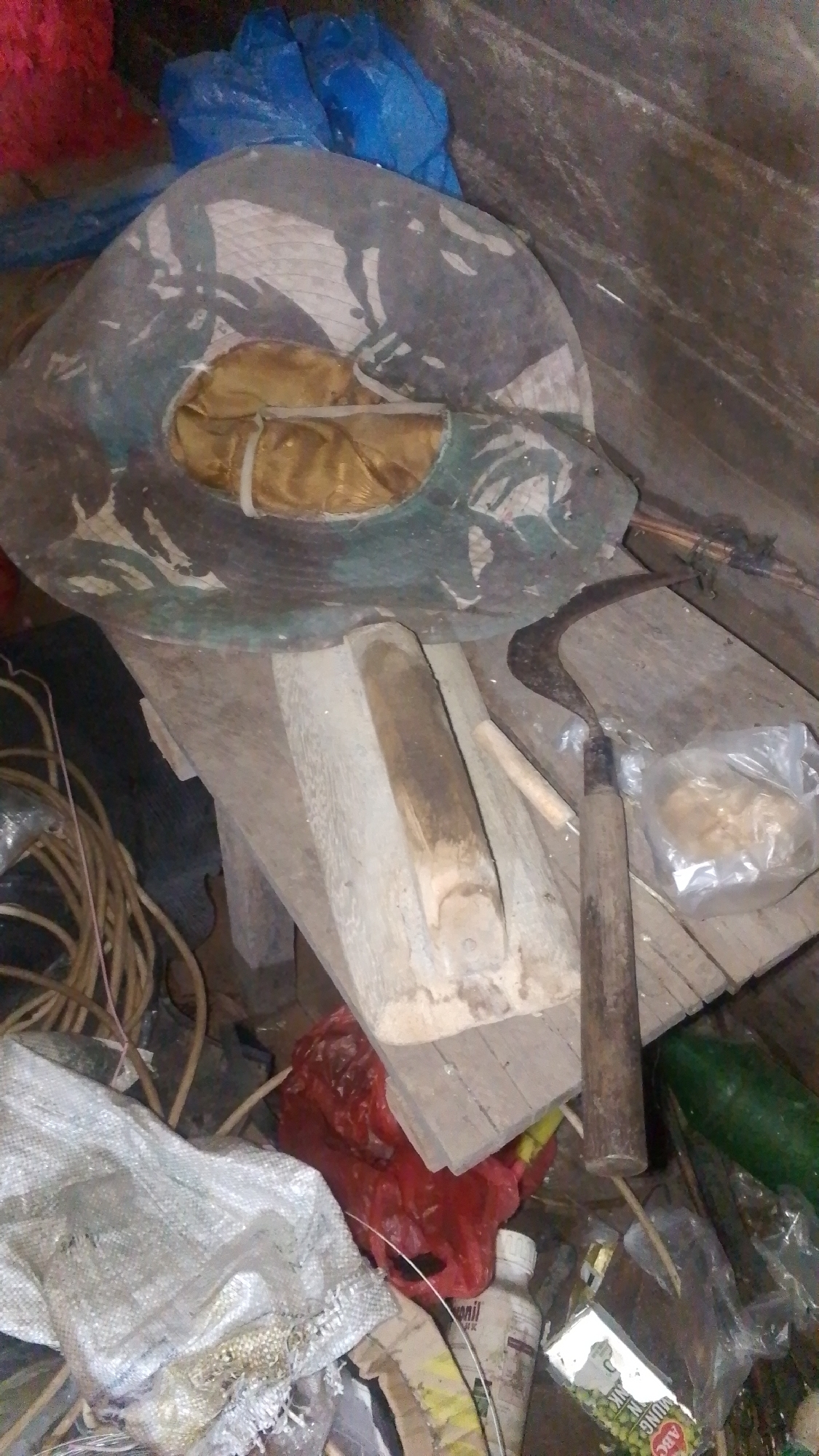 The image depicts a cluttered outdoor scene, possibly a storage area or a neglected corner. In the foreground, a large, circular object, resembling a hat or a woven tray with a patterned brim, is centrally positioned. It is made of a coarse material with distinct green and gray markings. Beneath this object is a rough wooden structure, possibly a stand or a partially constructed item, with visible grain and splinters.

To the right of the wooden structure, a sickle with a wooden handle lies on a wooden plank. The sickle appears old and somewhat rusted. Next to the sickle, a small plastic bag contains some light-colored granular material.

To the left of the main wooden structure, a bundle of thick, coiled ropes or cables is visible. Further to the left and towards the bottom, a torn white woven bag is present, with some debris scattered around it.

In the background and scattered throughout the scene are various other items. A large blue tarp or plastic sheeting is partially visible at the top, along with a bright red plastic bag. A long, dark green cylindrical object, resembling a large vegetable or a pipe, is situated on the right side of the image. A white plastic bottle with a label is also visible near the bottom center. A partially obscured package with visible lettering and green print is on the right side. The overall environment appears to be unorganized and possibly exposed to the elements, suggesting a utilitarian rather than a decorative space. There are no individuals present in the image, and no specific actions are depicted, beyond the static arrangement of objects.
