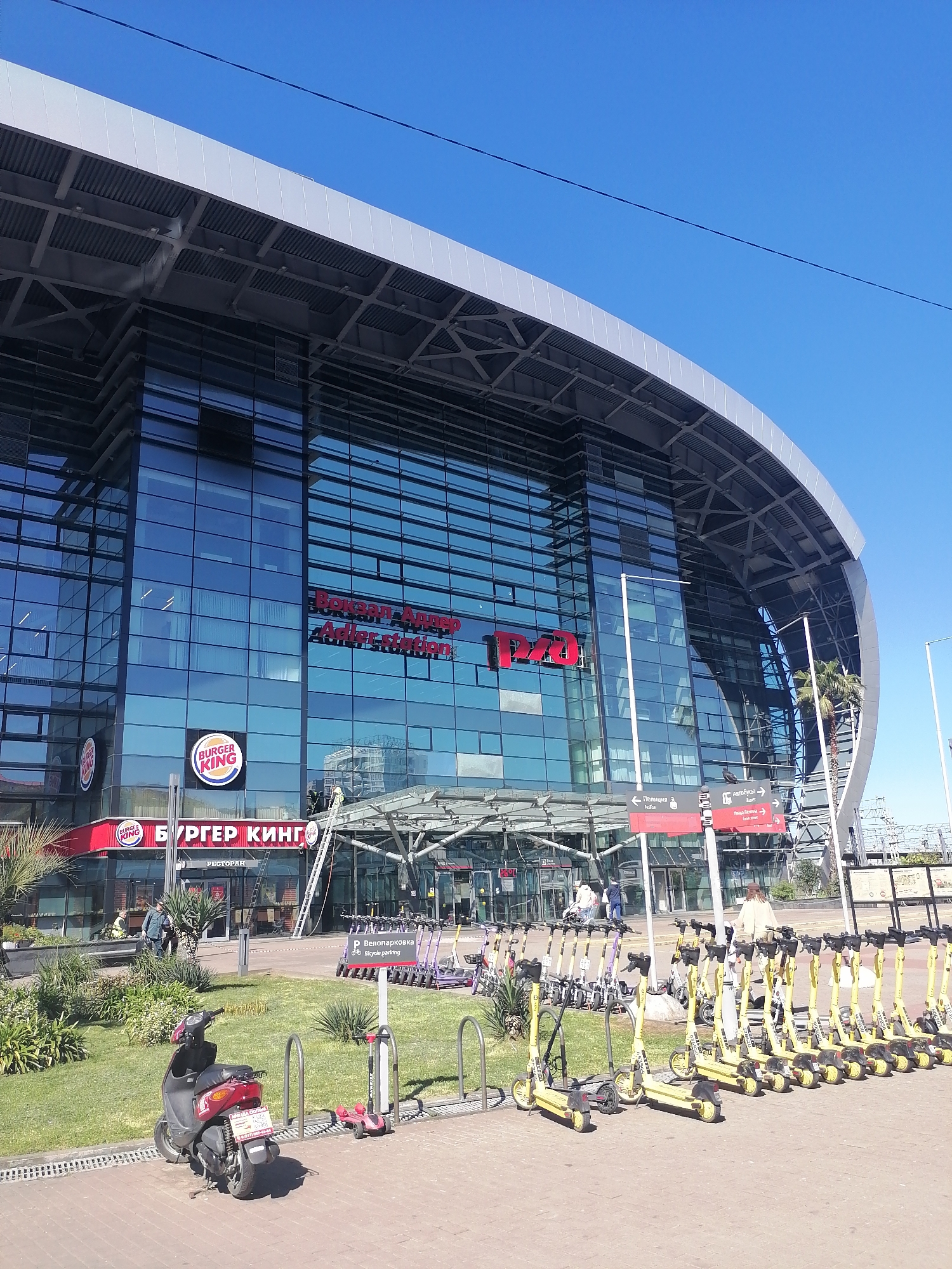 Here is a detailed description of the image:
The image shows a modern building with glass facade, identified as the Adler train station in Sochi, Russia ("Adler station" is written on the facade in both Russian and English).
On the ground level, to the left of the main entrance, there is a Burger King restaurant ("БУРГЕР КИНГ" is written on the facade in Russian). A person is on a ladder near the Burger King sign, and two other people are seen near the restaurant entrance.
In front of the building, a parking area is visible with numerous electric scooters. To the left, there is a parking spot with a parked motor scooter. A sign indicates "Р Велопарковка Bicycle parking." Two directional signs are also visible, one directing towards "Полиция" (police) and another towards "Автобусы" (buses).
The sky is clear and blue, suggesting a sunny day.