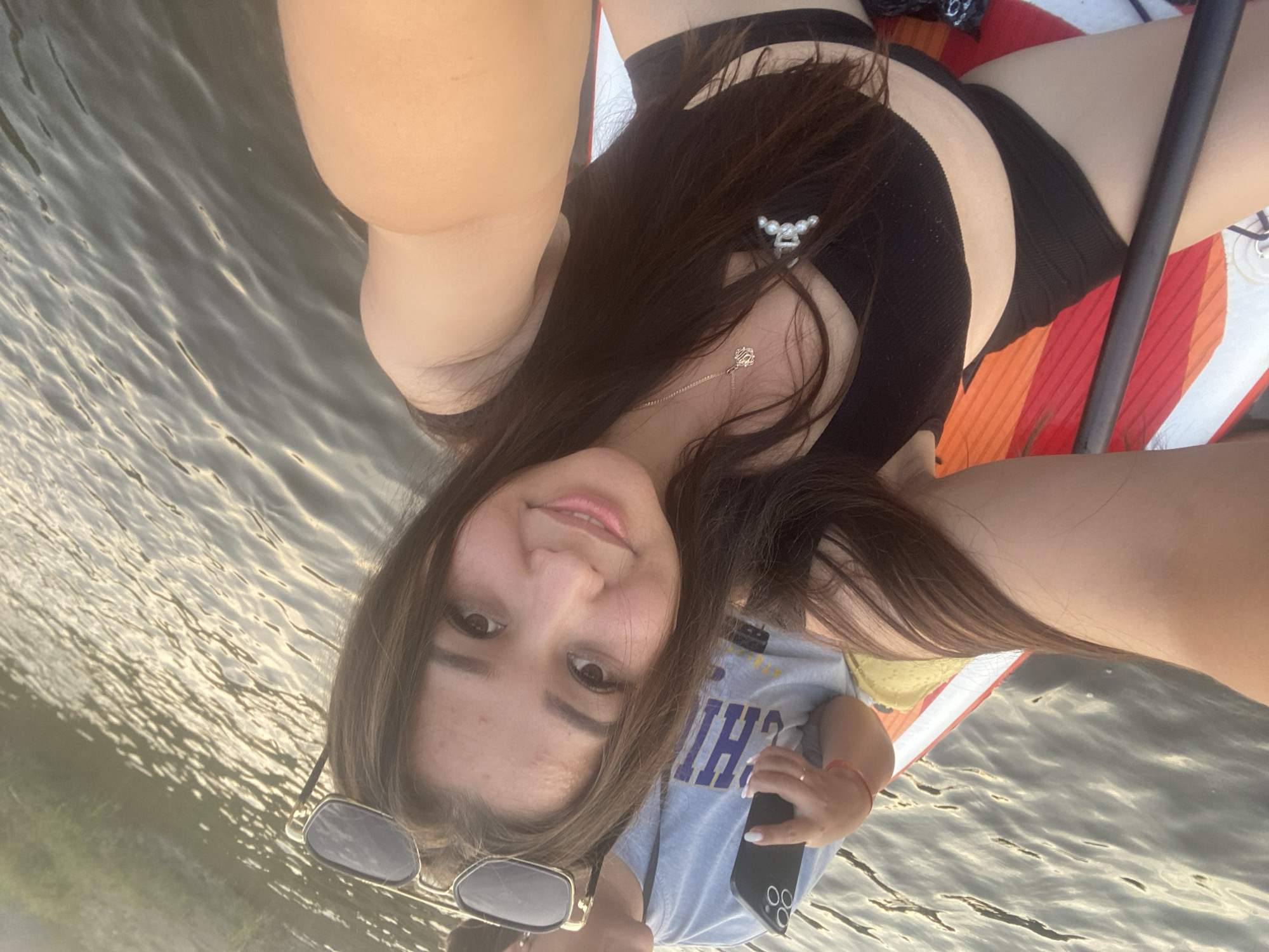 A young woman is taking a selfie while sitting on a paddleboard in the water. She is wearing a black bikini top and black bottoms, with a silver necklace and a small pearl embellishment on her top. She has long, dark brown hair and is wearing sunglasses. Her face is framed by her hair, and she is smiling gently at the camera. The paddleboard has a colorful design with red, orange, and white stripes.
In the background, the water is calm with gentle ripples reflecting the sunlight, suggesting it is daytime, likely late afternoon given the warm light. The woman is holding her phone in her right hand, likely to take the selfie. On her left arm, she is wearing a thin red bracelet. She also has a ring on her right hand.
The image is taken at a slightly upward angle, showing the woman from the chest up, with her legs and part of the paddleboard extending out of frame. The scene is peaceful and relaxed, indicating a leisurely activity. The text "CHI" is visible on the t-shirt of another person partially visible in the lower left of the frame. The overall mood is casual and summery.