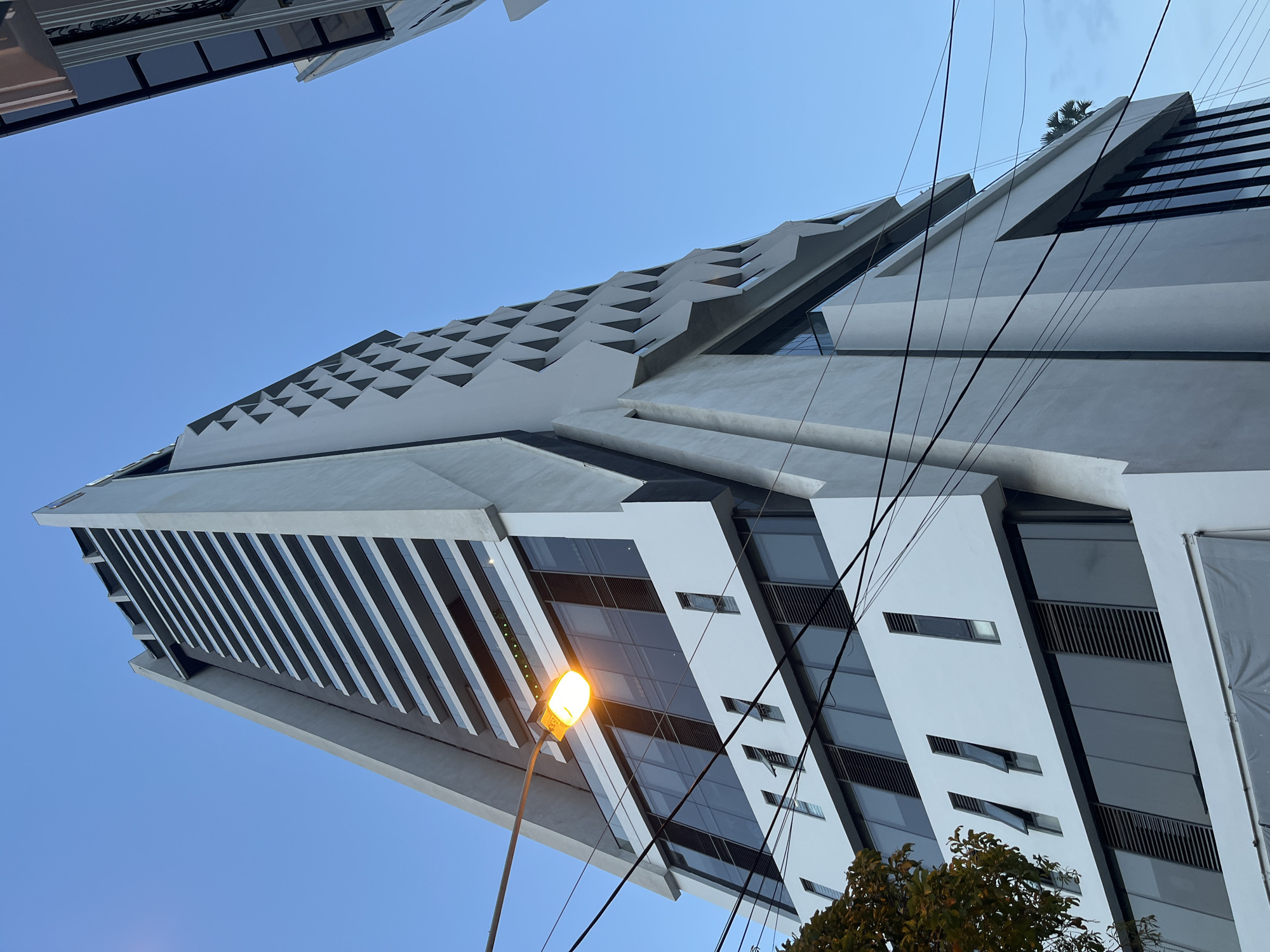The image is a low-angle, full-shot photograph of a modern, multi-story building taken in the city of Nha Trang, Vietnam. The building, predominantly white with large dark-tinted windows, features a unique architectural design with geometric patterns of triangular cutouts on its upper facade. The sky is clear and blue, indicating daytime. Several black electrical wires are visible in the foreground, crisscrossing the frame and obscuring parts of the building's lower levels. A street lamp with a warm orange glow is positioned to the left of the building, partially illuminated. The bottom of the image shows what appears to be construction or renovation materials covered by a gray tarp. A tree with sparse, autumnal foliage is visible on the far left. The overall scene suggests an urban environment, with the building's striking design contrasting with the functional presence of overhead utility lines.